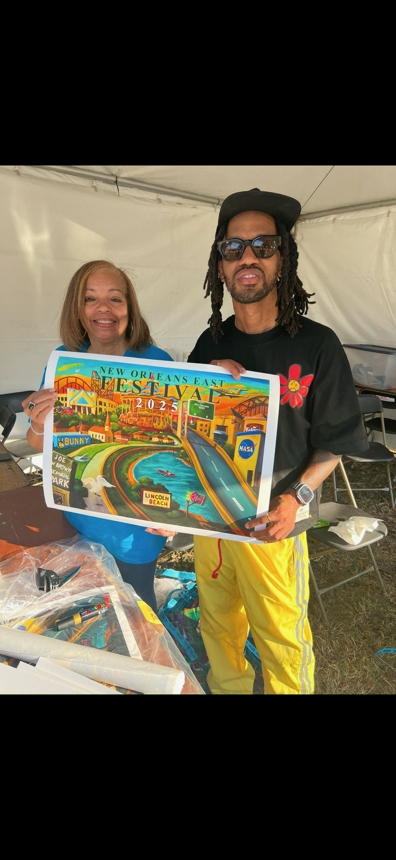 Two people holding a colorful illustrated poster for the New Orleans East Festival 2023. The woman on the left is smiling, wearing a blue shirt, and the man on the right is wearing sunglasses, a black cap, a black T-shirt with a red flower emoji, and