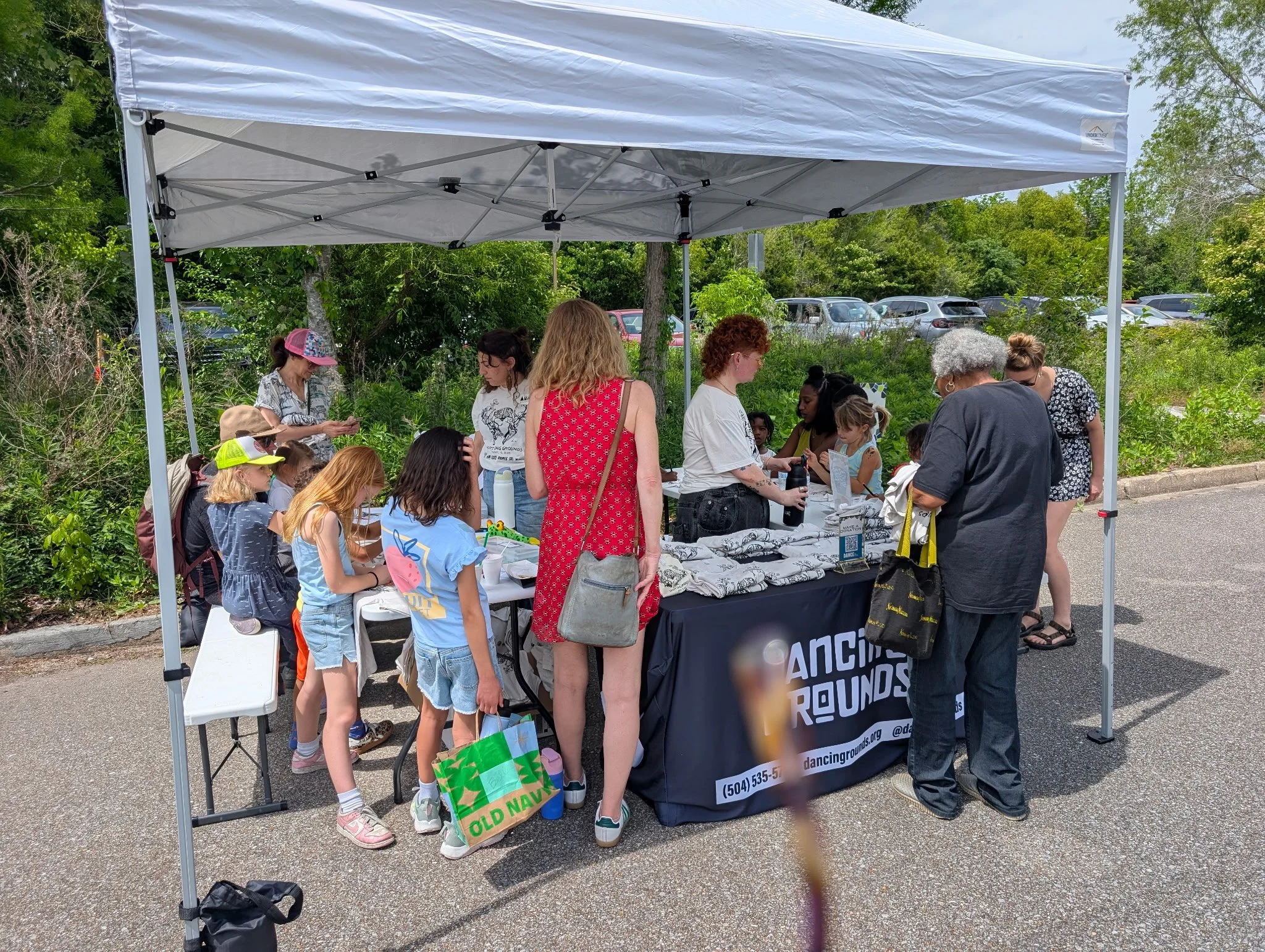 People gather under a white canopy tent at an outdoor event. Some are standing, others sitting at a table with items for sale or display. There are trees and parked cars in the background.