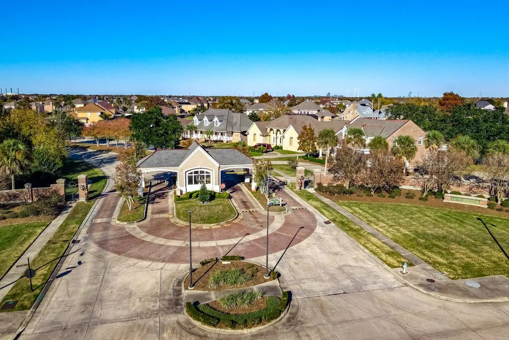 Aerial view of a suburban neighborhood with a gated community entrance, houses, green lawns, trees, and a clear blue sky.