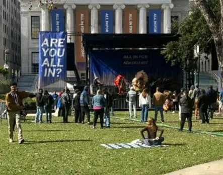 People gathered on a grassy area outside a building with banners, watching a stage performance with a person speaking or acting, and some individuals sitting on the grass.