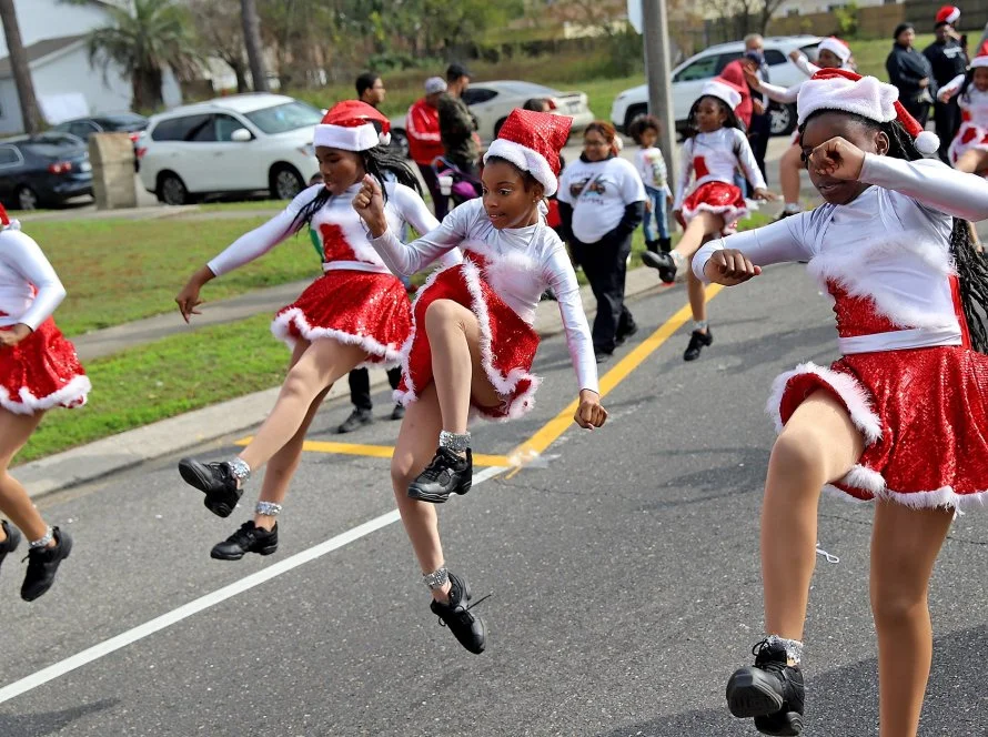 Children in festive Christmas costumes dancing and jumping in a parade on the street, wearing red and white outfits with Santa hats.