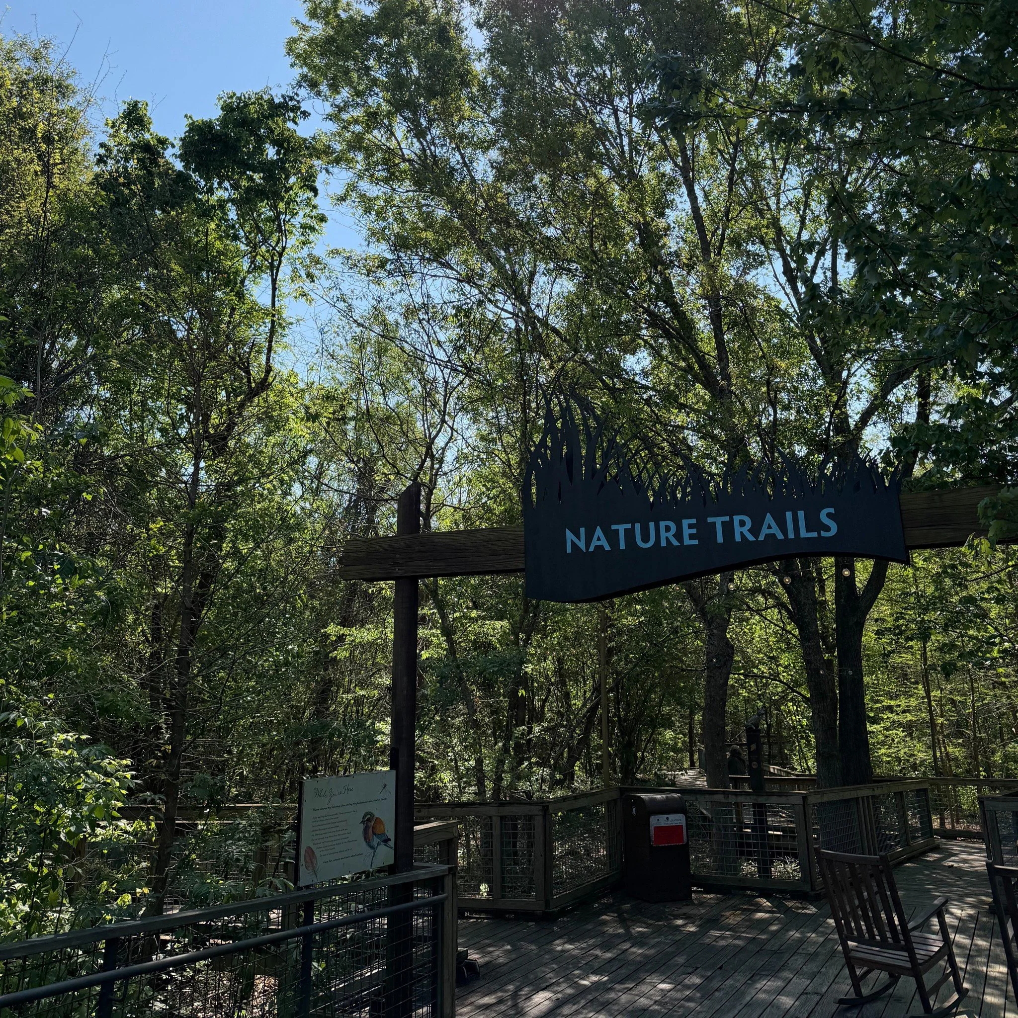 Entrance to a nature trail area with trees overhead, a sign that reads 'Nature Trails,' and a wooden deck with benches and informational panels.