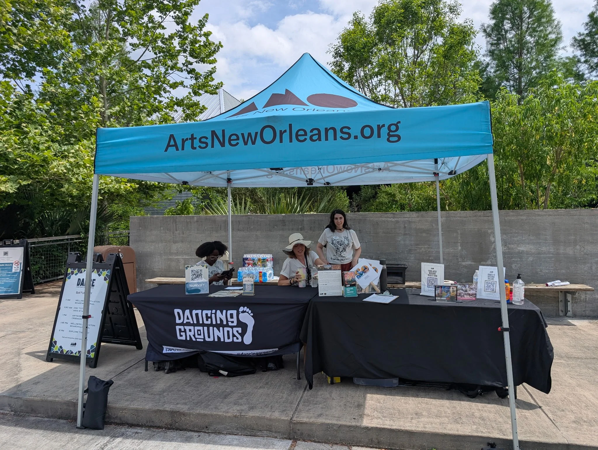 A blue tent with "ArtsNewOrleans.org" printed on it. Below is a table with plenty of pamphlets and QR codes, some people sitting at the table. In front, a black tablecloth with white text reading "Dancing Grounds," with a footprint logo. A woman and 