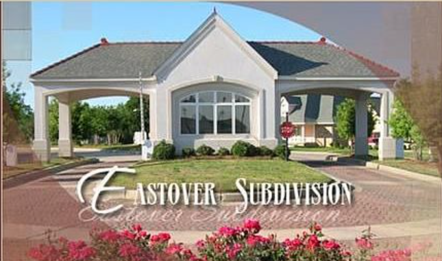 Entrance of E-Estover Subdivision with a white building, brick driveway, and pink flowers in front.