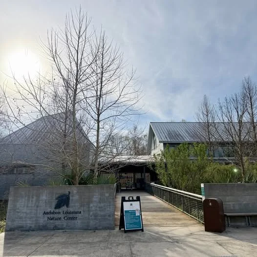 Entrance to Audubon Louisiana Nature Center with a concrete sign, leafless trees, a walkway, and a building with a sloped metal roof.