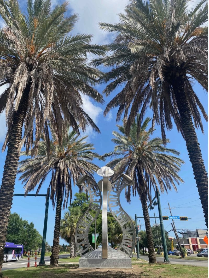 A metal public art sculpture featuring a diamond shape with cut-out patterns, surrounded by tall palm trees against a blue sky with some clouds, near a street with traffic lights and buildings.