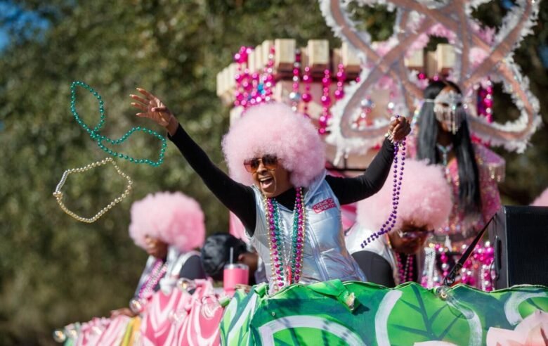 Person with pink afro wig and sunglasses dancing and holding necklaces during a parade float decorated with elaborate pink and green designs and a large octopus sculpture in the background.