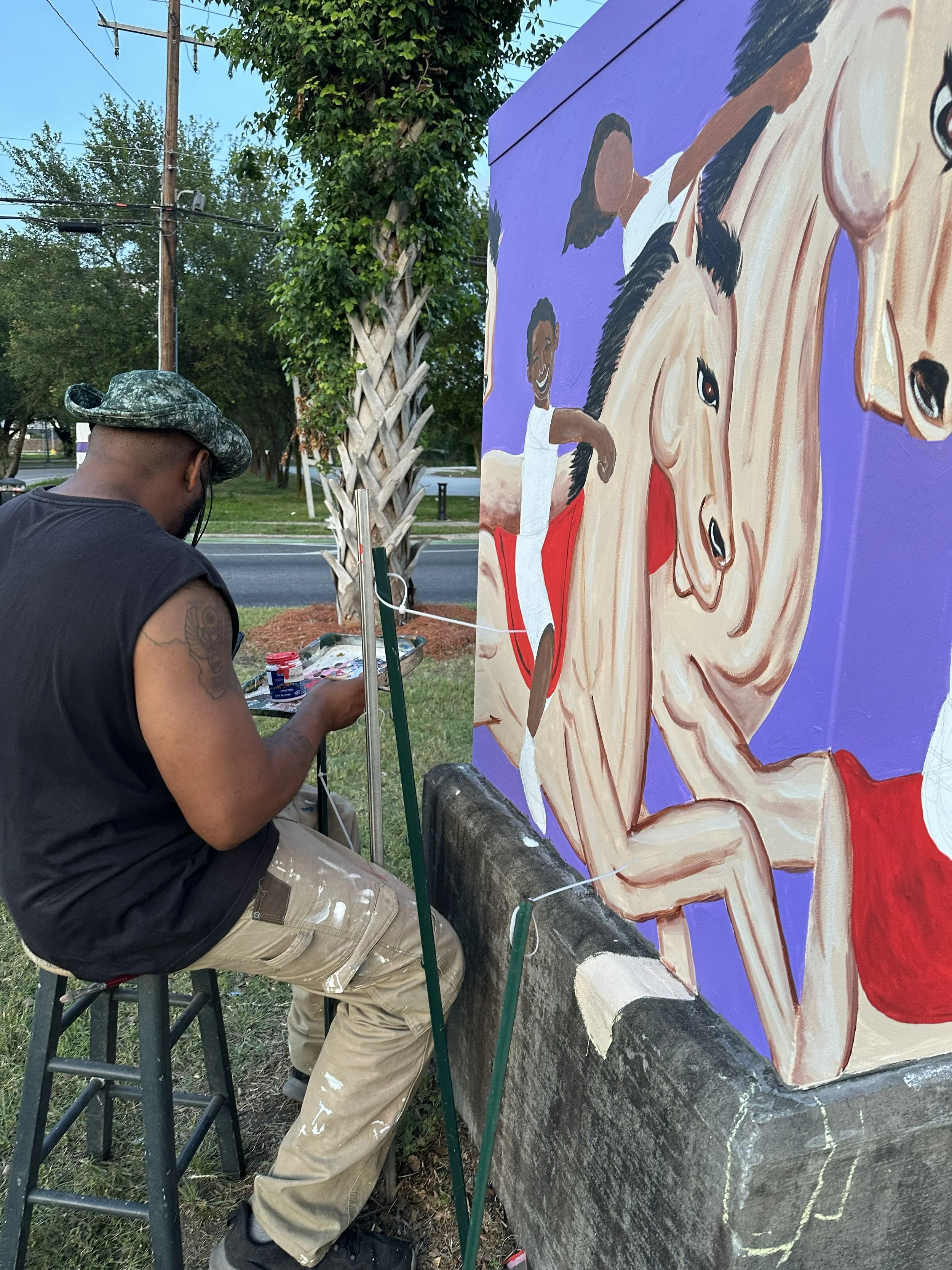 An artist painting a colorful mural of a smiling person with dark hair and a horse on a large purple canvas outdoors. The artist is seated on a stool, wearing a camouflage hat and beige pants.