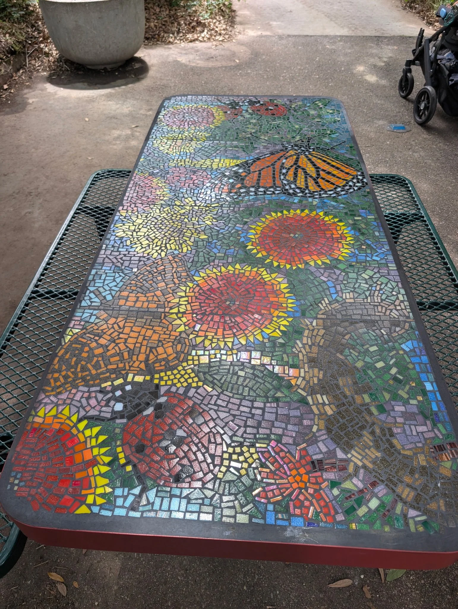 A decorated mosaic table with colorful patterns, including butterflies, flowers, and geometric shapes, situated outdoors on a paved surface.