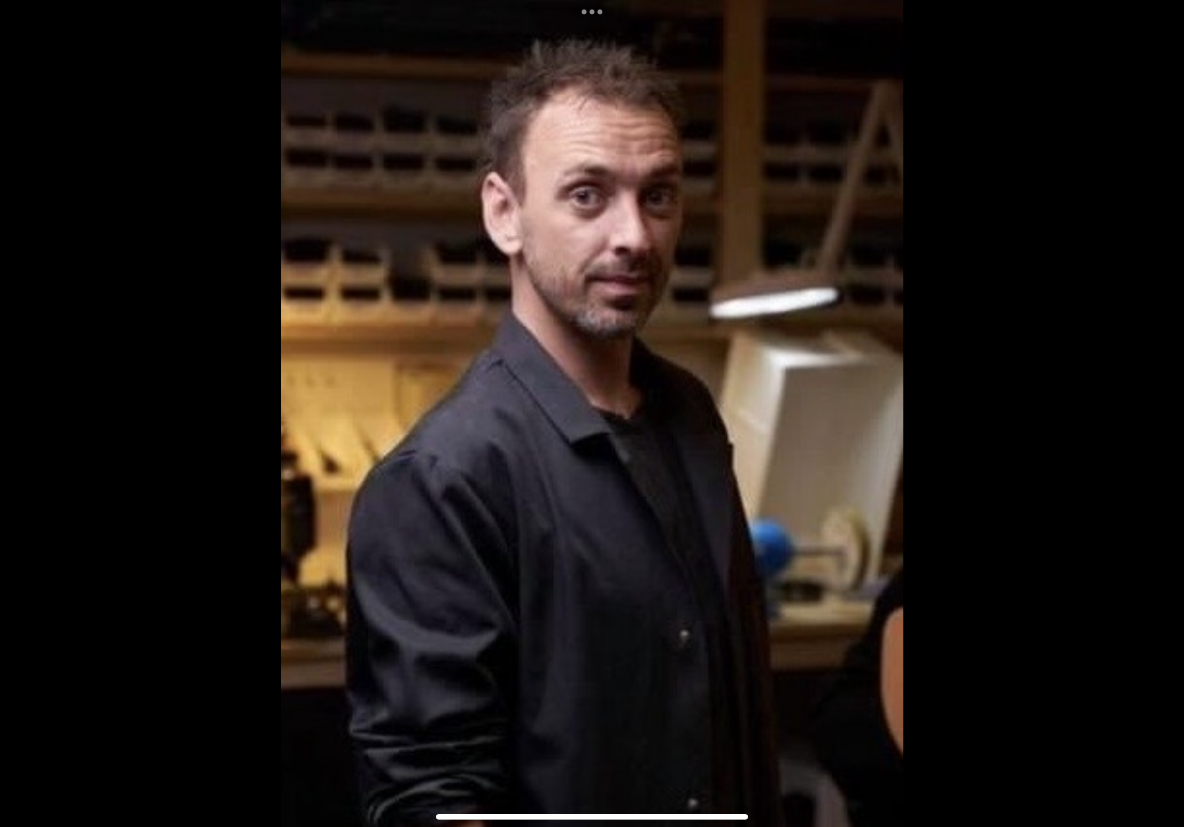 A man with short, dark, slightly spiked hair and light facial hair, wearing a black shirt, standing in a woodworking shop with woodwork and tools in the background.