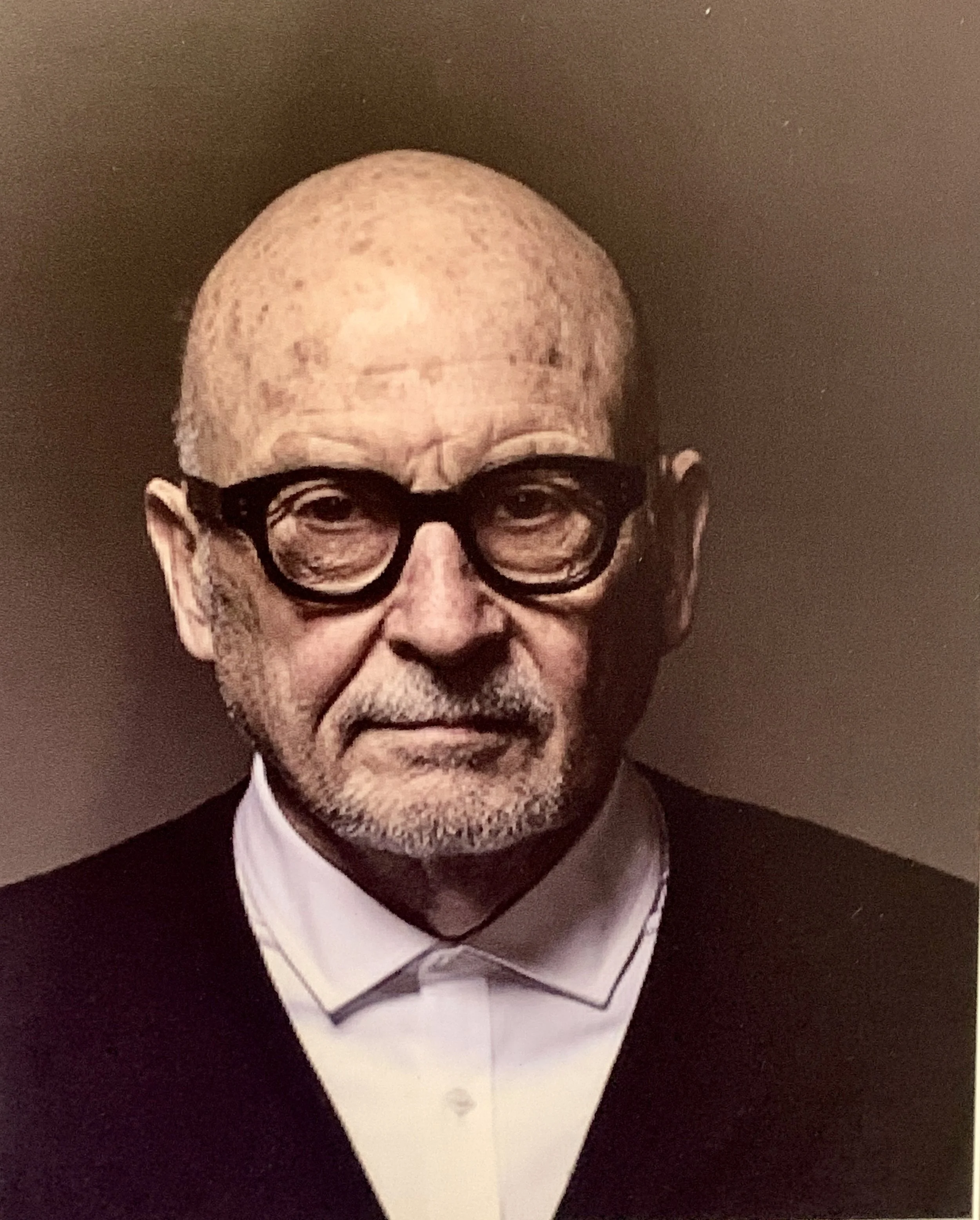 A portrait of an elderly man with a bald head, wearing large round black glasses, a white shirt, and a dark suit.