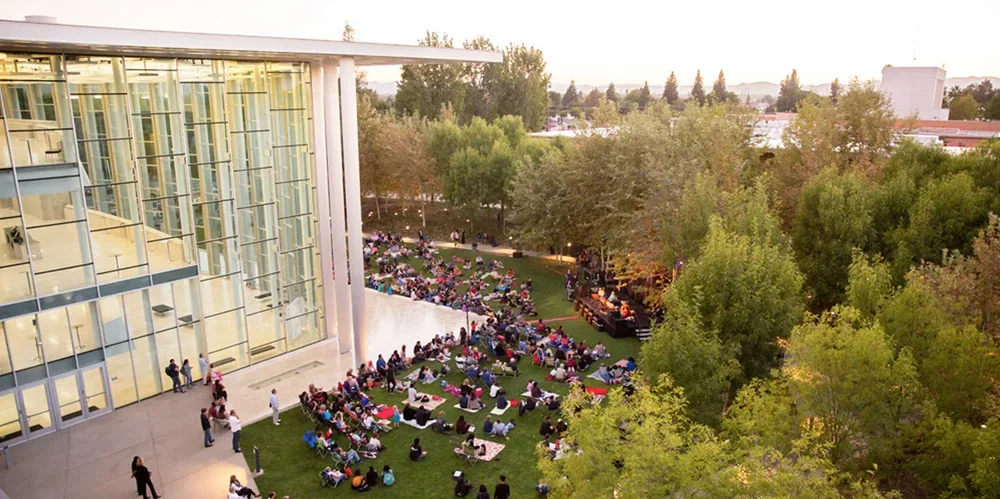 Large outdoor gathering of people sitting on the grass and under trees near a modern building with tall glass windows, possibly attending an event or concert in the late afternoon or early evening.