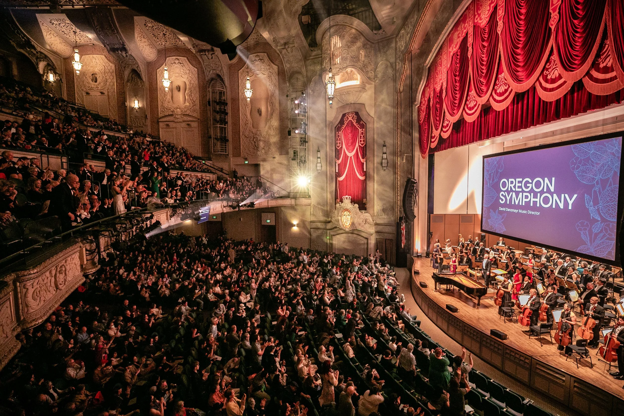 Orchestra performing in a grand theater with red curtains and an audience clapping