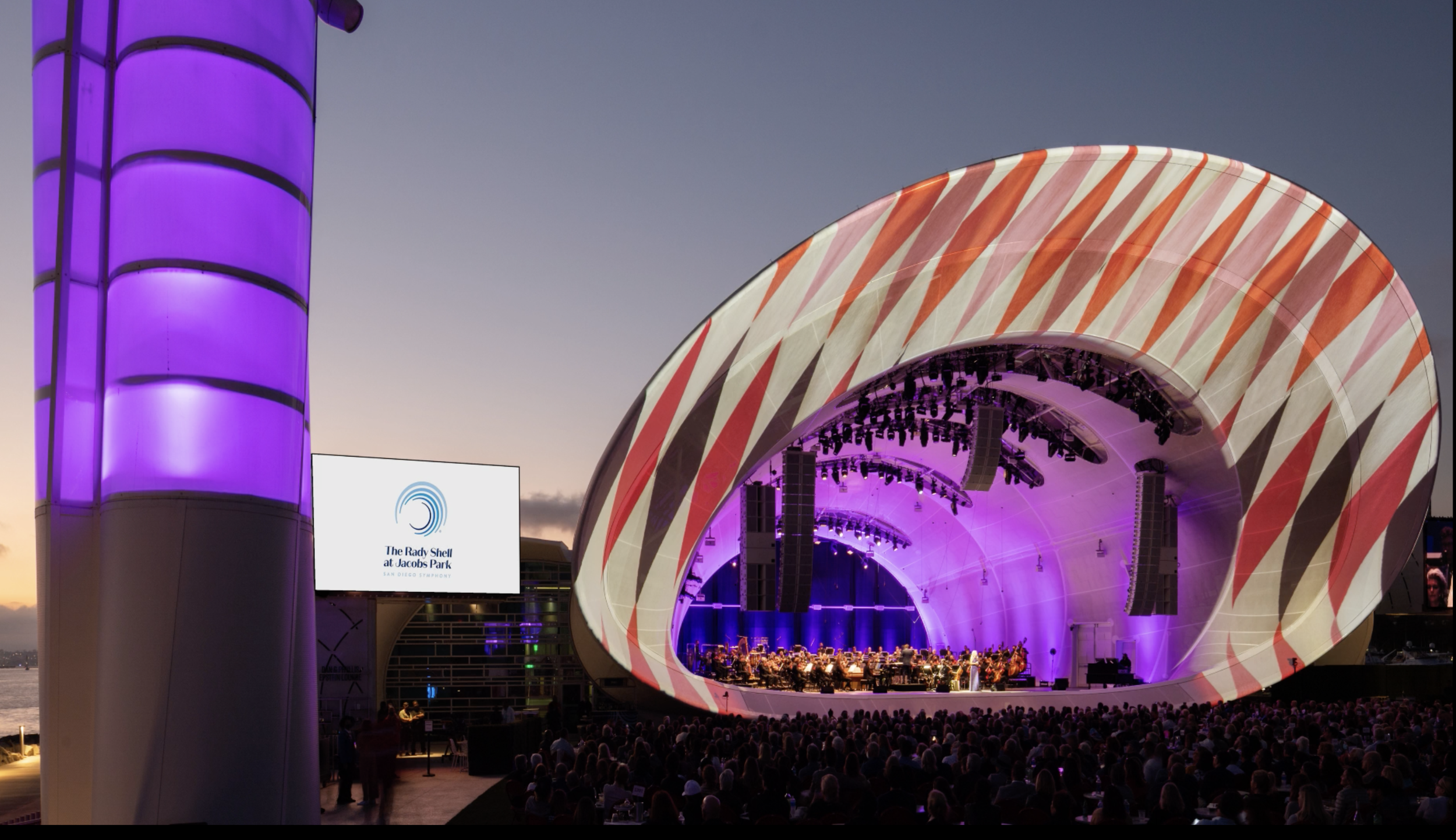Outdoor concert stage at sunset with purple lighting, modern shell-shaped design, and a large audience seated in front. A screen displays "The Rady Shell at Jacobs Park" logo.