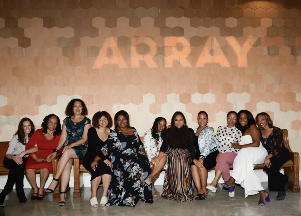 A group of nine women sitting on a bench, smiling at the camera, celebrating the opening of a new theater.