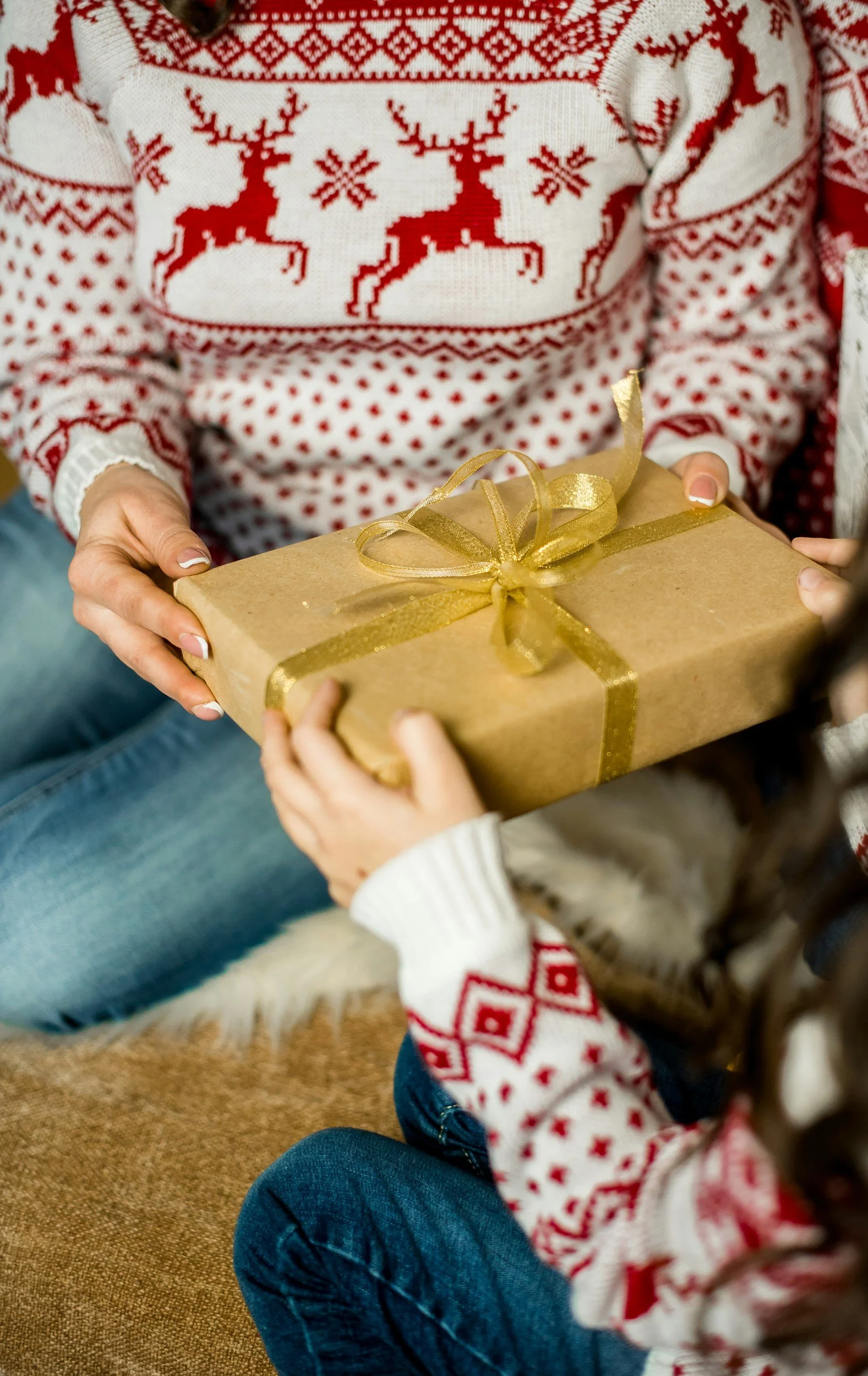 Person in a festive red and white Christmas sweater giving a wrapped gift with a gold ribbon to a child wearing a matching sweater.