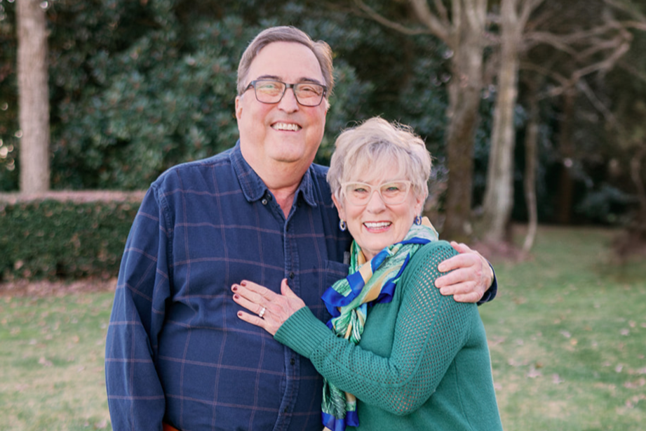 Smiling elderly couple embracing outdoors in a park with trees and grass in the background.