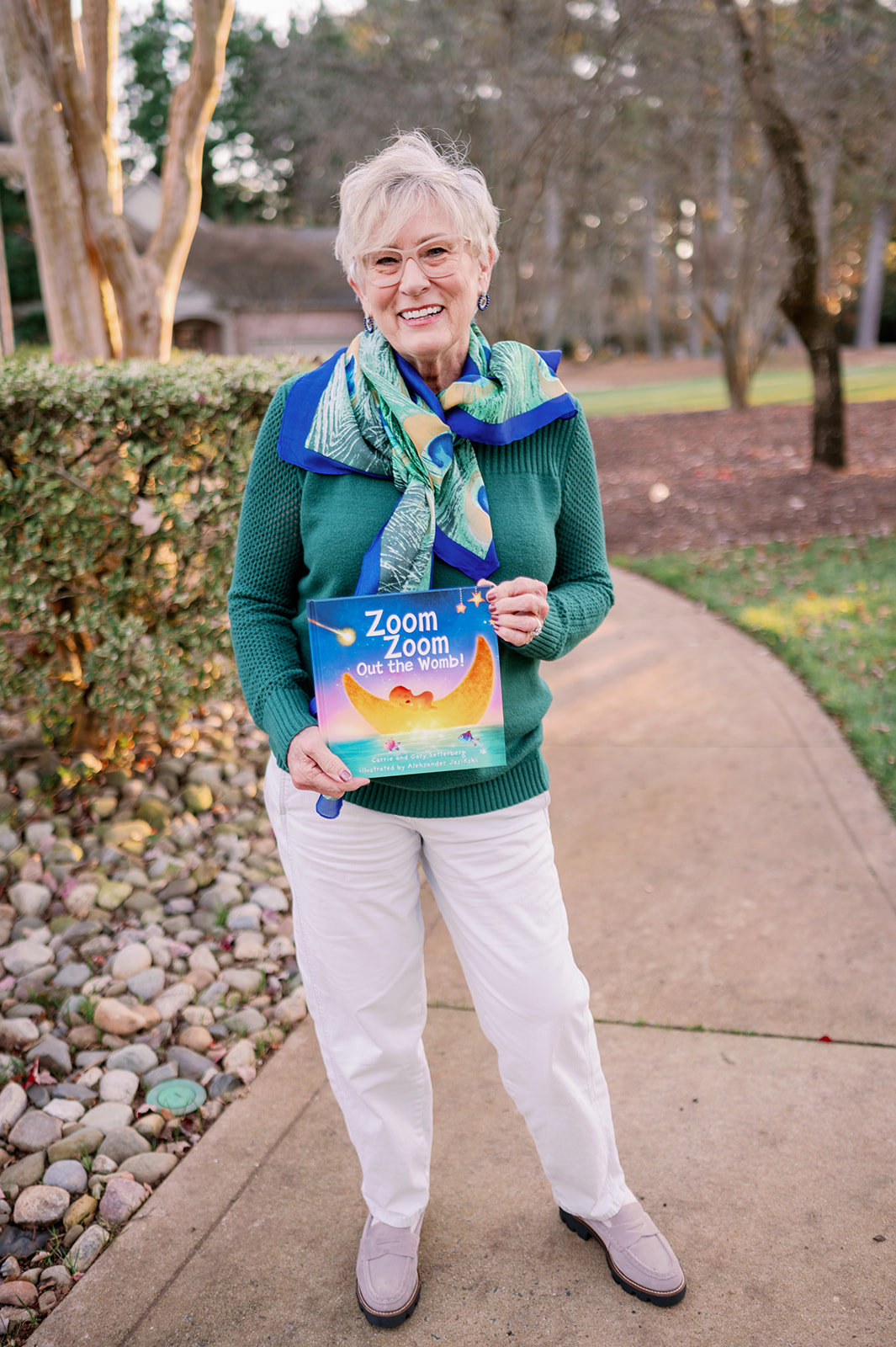 An older woman standing outdoors on a sidewalk holding a children's book titled 'Zoom Zoom Out the Womb!' She is smiling and wearing glasses, a green sweater, white pants, gray shoes, and a colorful scarf. The background includes trees, bushes, and a house.