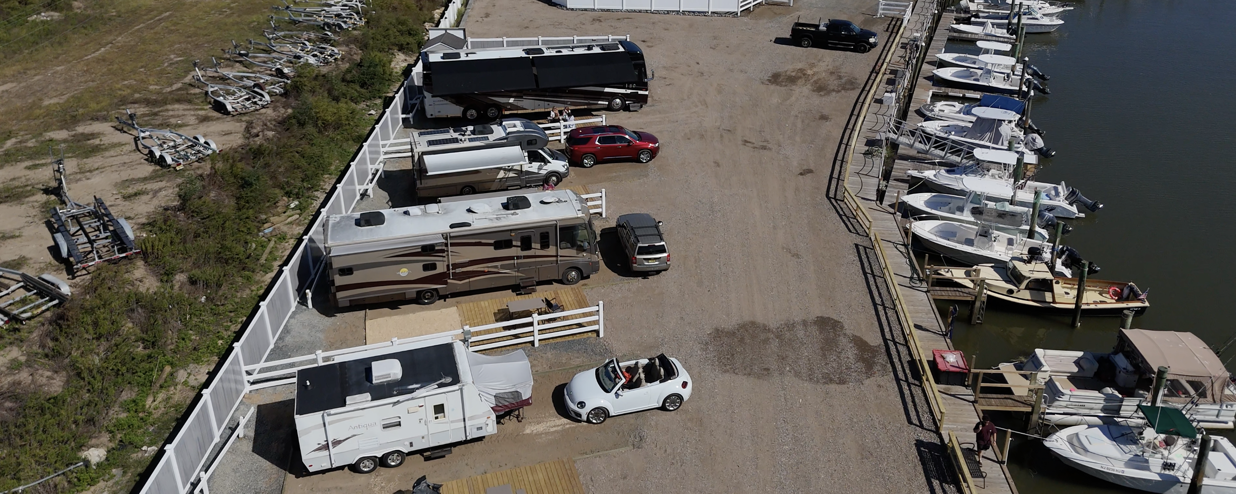 An aerial view of a marina parking lot with boats docked on the water, and various vehicles, including RVs, cars, and trucks, parked on land divided by a white fence and a gravel road.