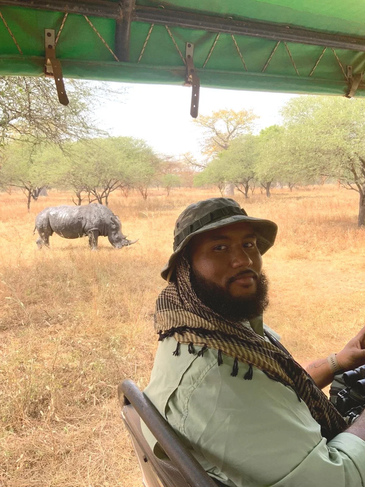 Kareem sits in an open vehicle, looking toward the camera and wears a green shirt, patterned scarf, and green bucket hat  Behind him, a rhinoceros grazes in a dry, grassy landscape with scattered trees.
