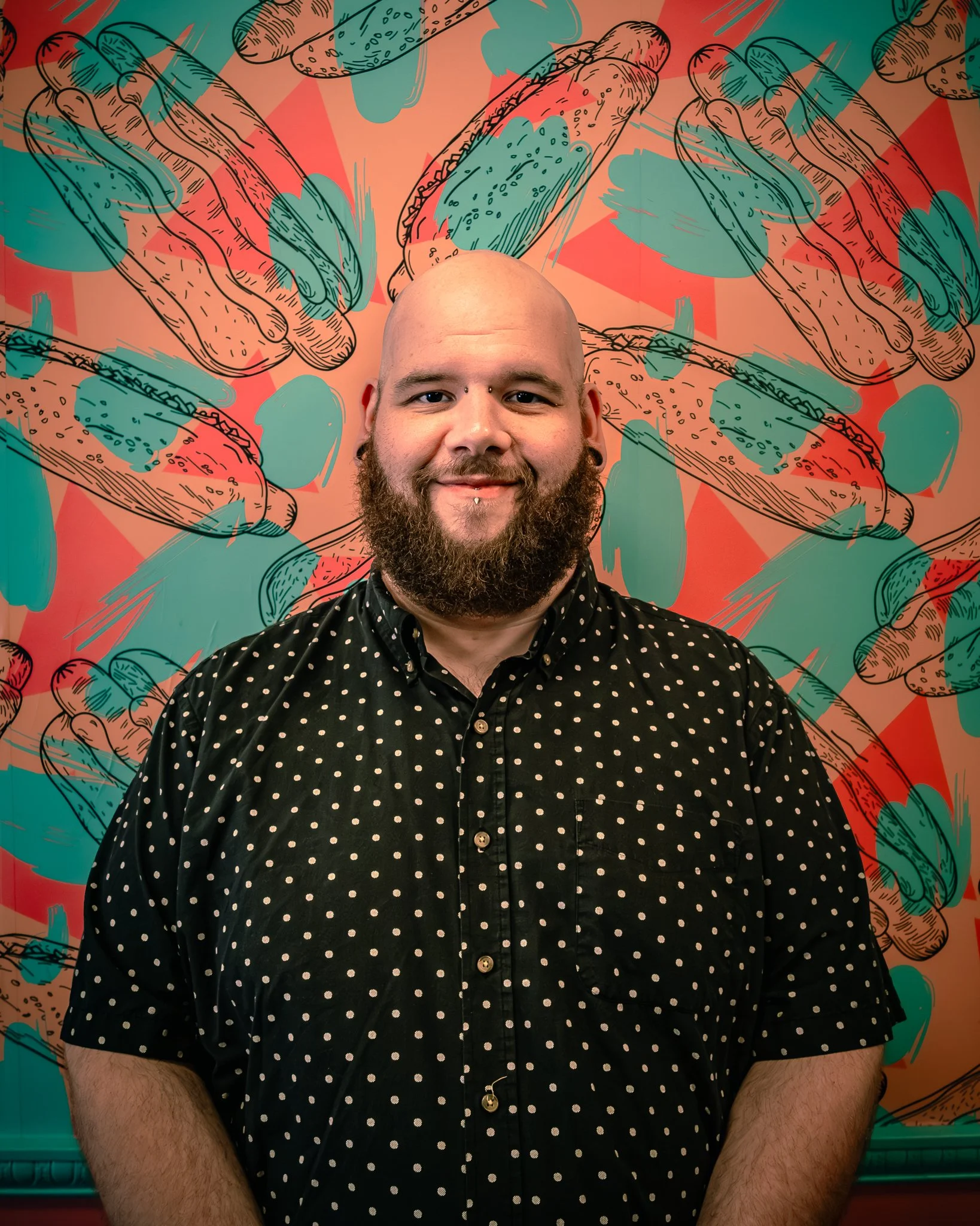 A man with a beard and piercings smiling, standing in front of a colorful, comic-style mural featuring hot dogs and abstract shapes.