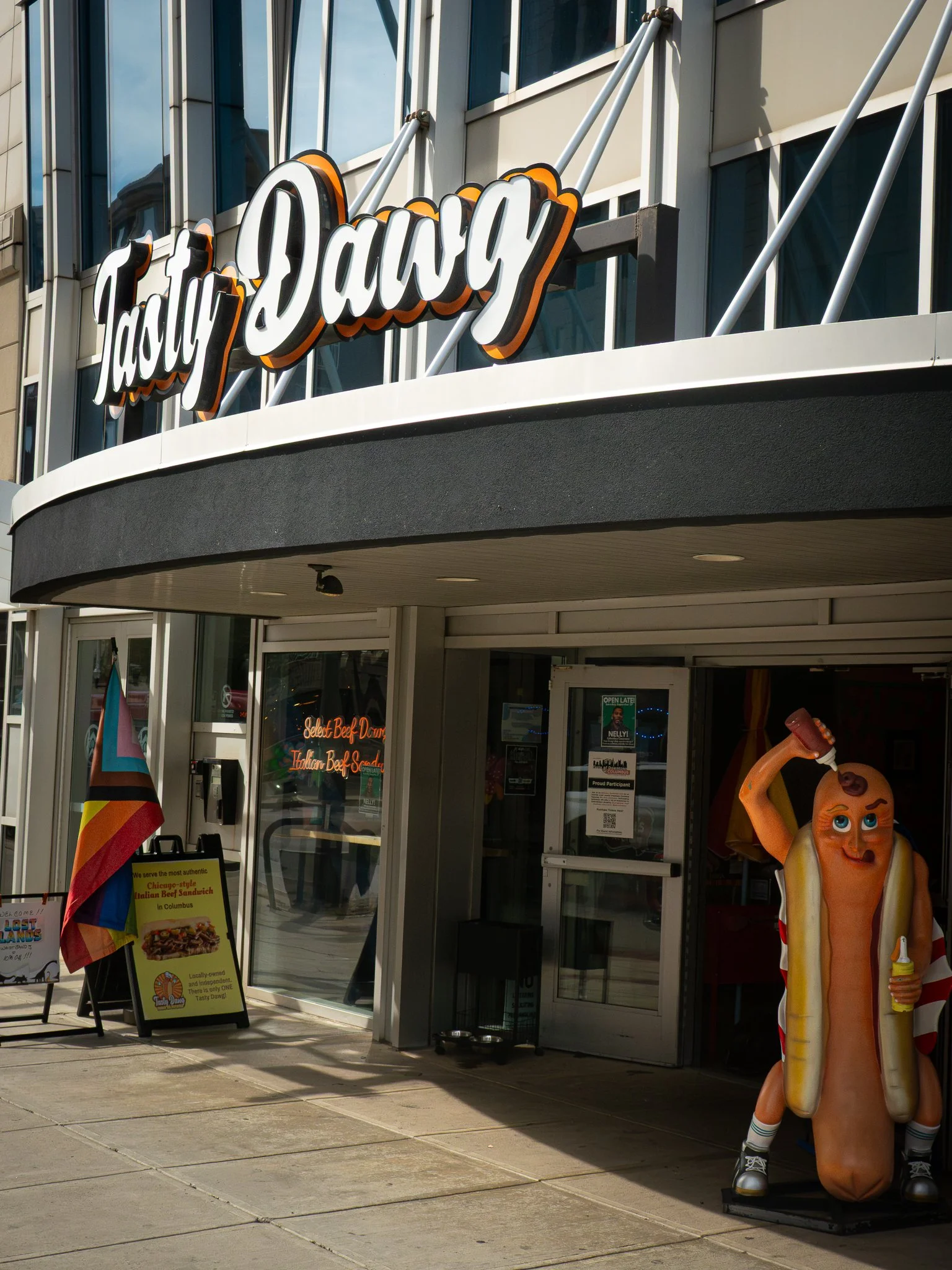 Facade of Tasty Dawg restaurant with a hot dog mascot wearing sneakers and sunglasses, holding a ketchup bottle and a yellow ice cream cone, standing outside on the sidewalk.
