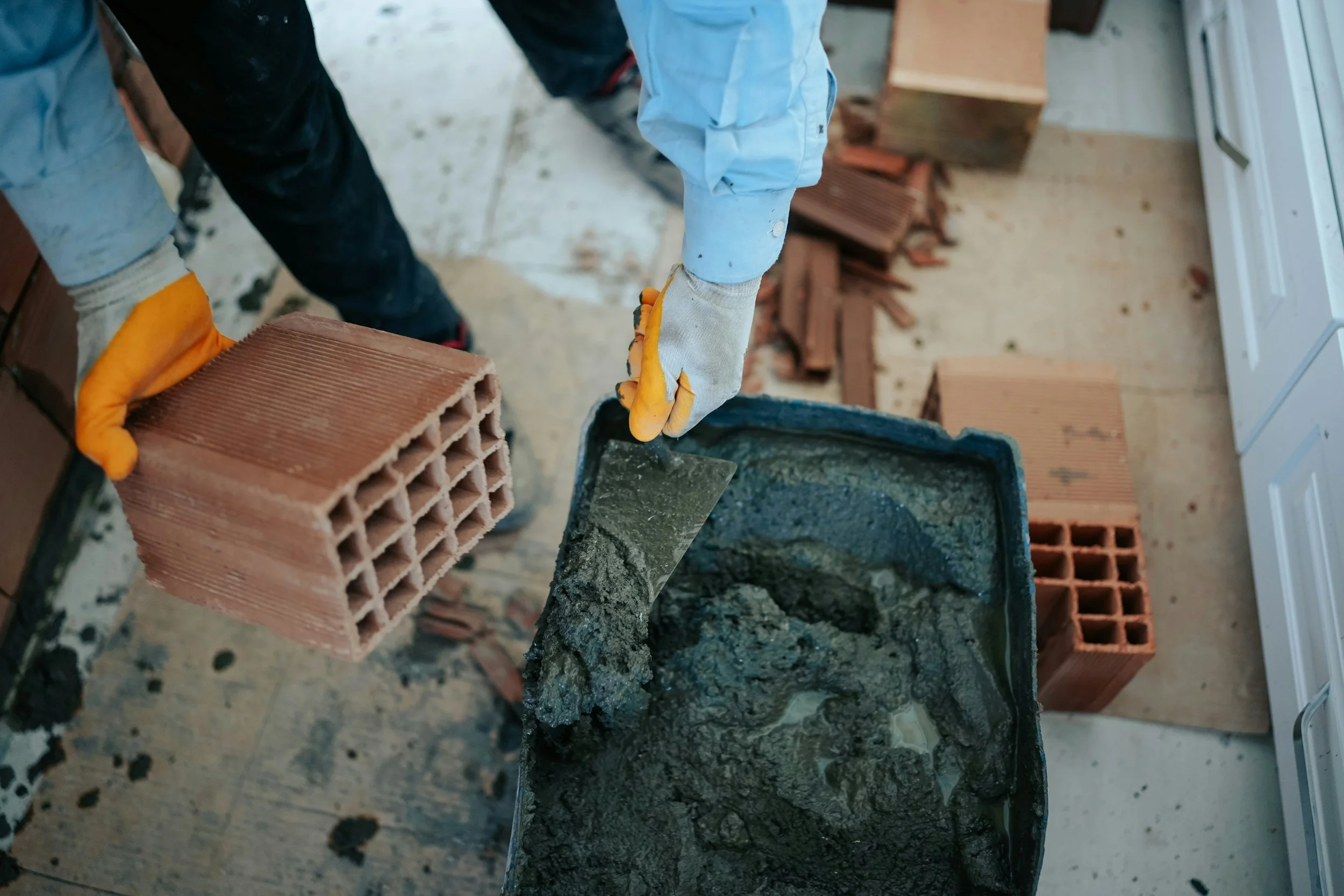 Person mixing mortar with a trowel while holding a brick, wearing gloves and work clothes, on a construction site with bricks and a concrete surface.