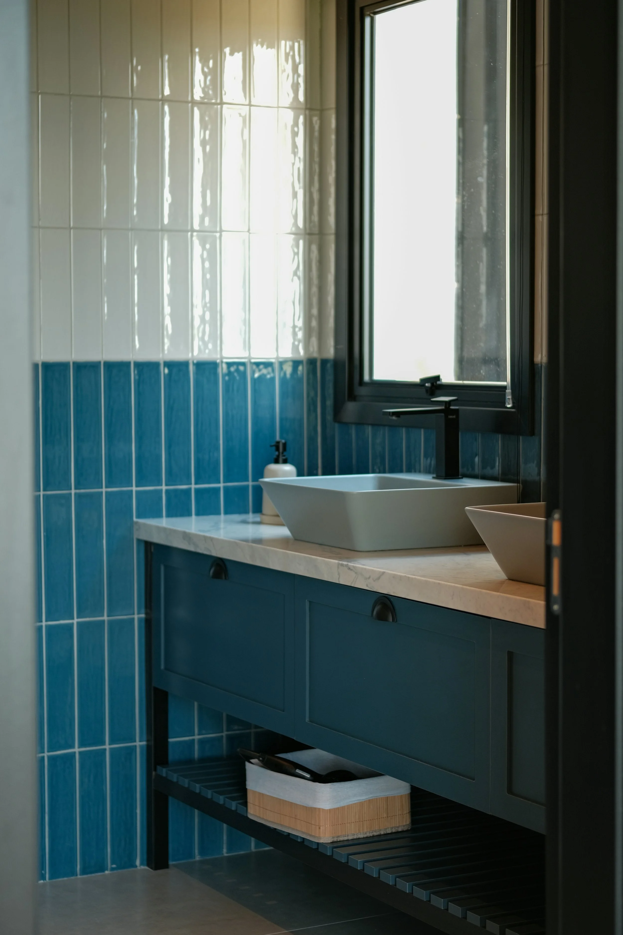 A modern bathroom with white and blue tiled walls, a black-framed window, a white rectangular vessel sink with a black faucet, and a countertop with a soap dispenser. There is a blue cabinet with black handles underneath, and a black slatted shelf holding a box.