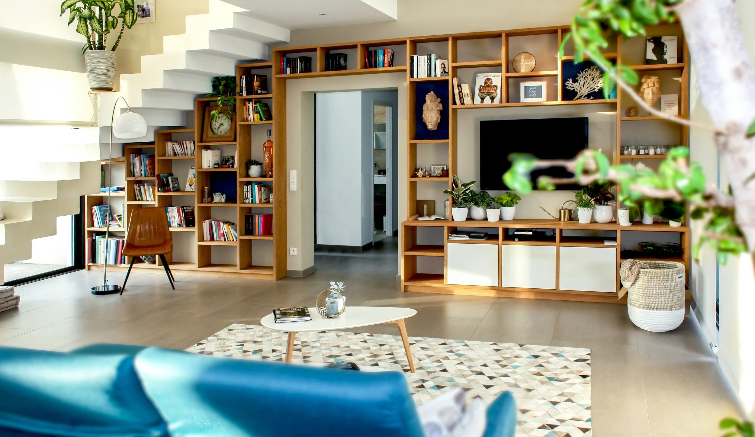 Modern living room with a wooden shelving unit, a TV, indoor plants, a brown chair, a floor lamp, a patterned rug, and a small white coffee table.