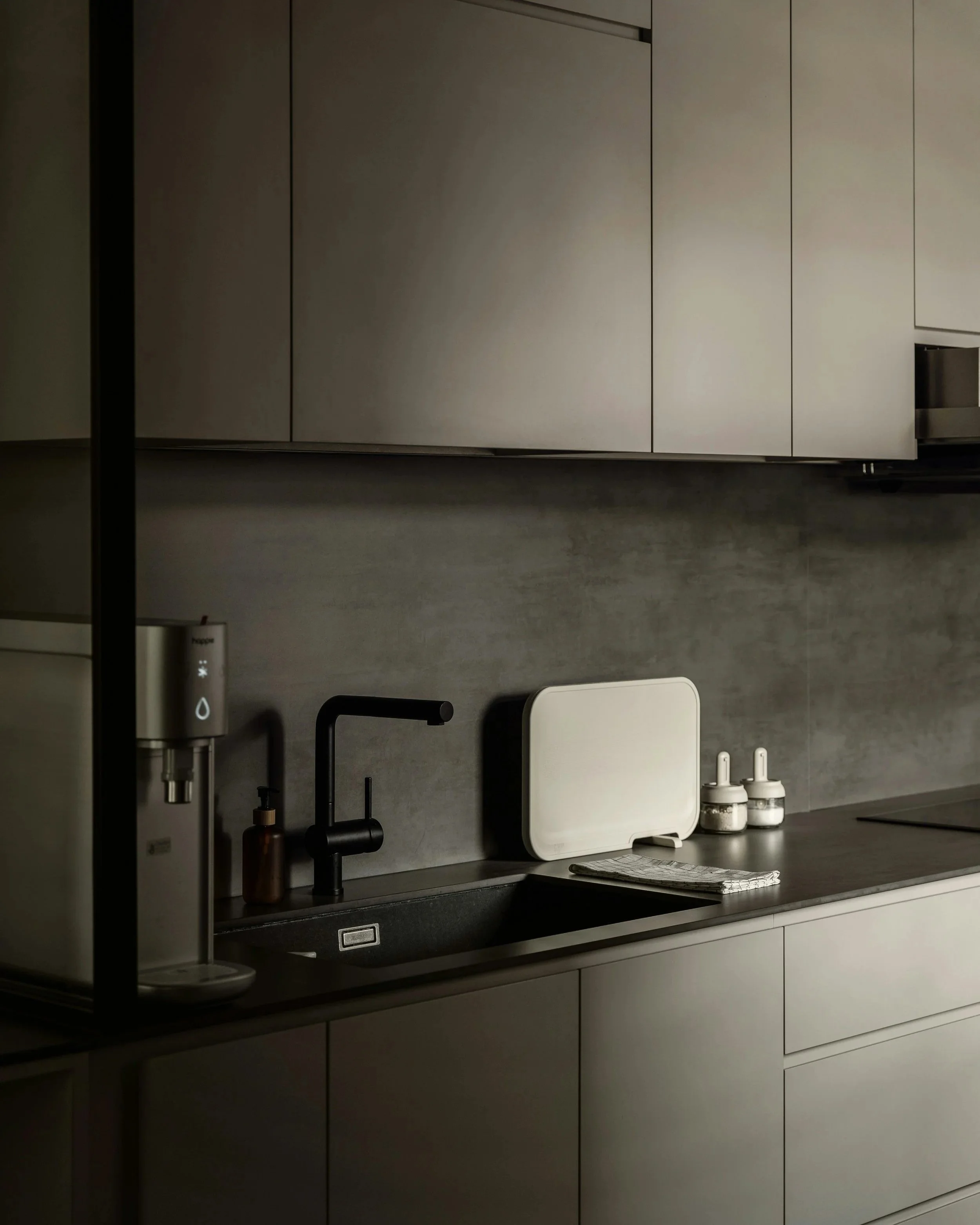 A modern kitchen with gray cabinets, a dark countertop, a black faucet, a white dish drying mat, two small spice jars, and a white water filter machine.