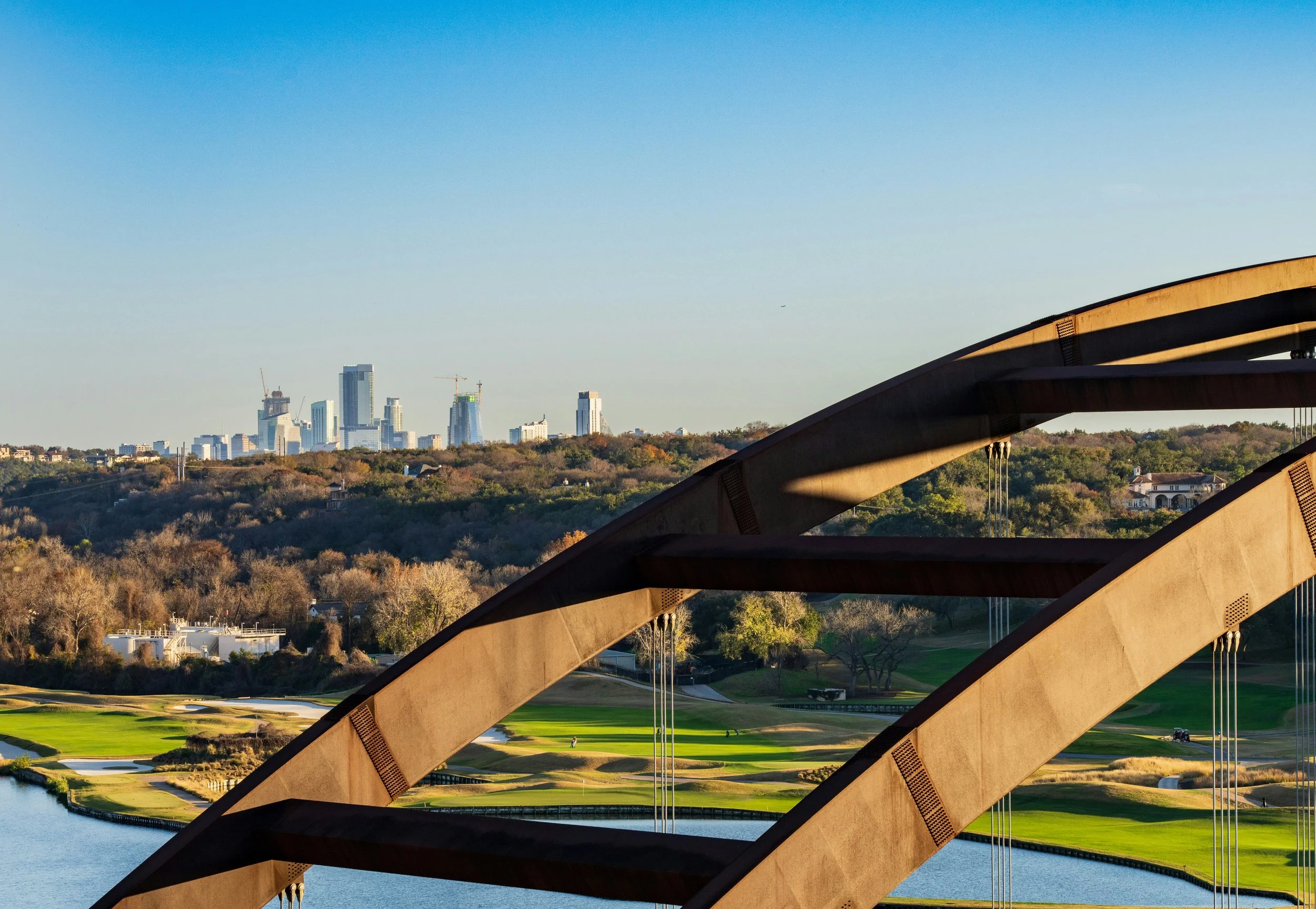 View of a city skyline with tall buildings in the distance, viewed through the arches of a bridge overlooking a golf course with water hazards.