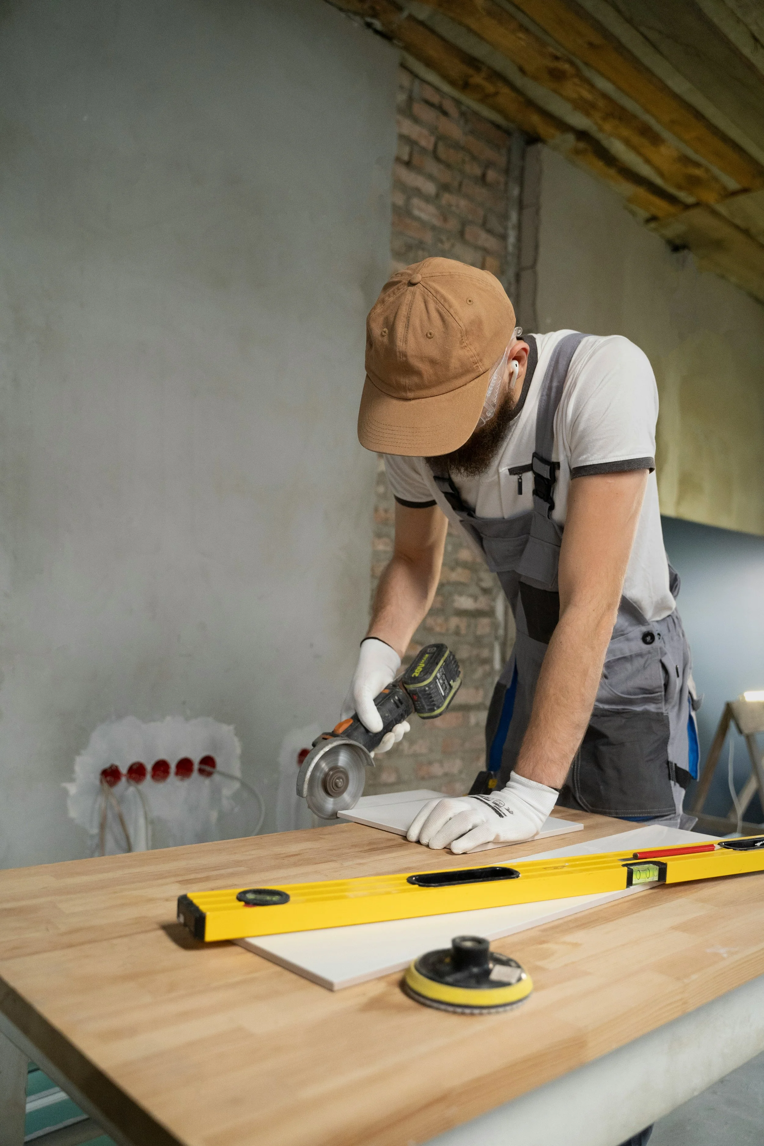 A man wearing a brown hat, white T-shirt, and gray overalls using a cordless drill to cut or drill a piece of wood on a workbench. There are tools like a level and a sander on the workbench in a room with unfinished walls and ceiling.