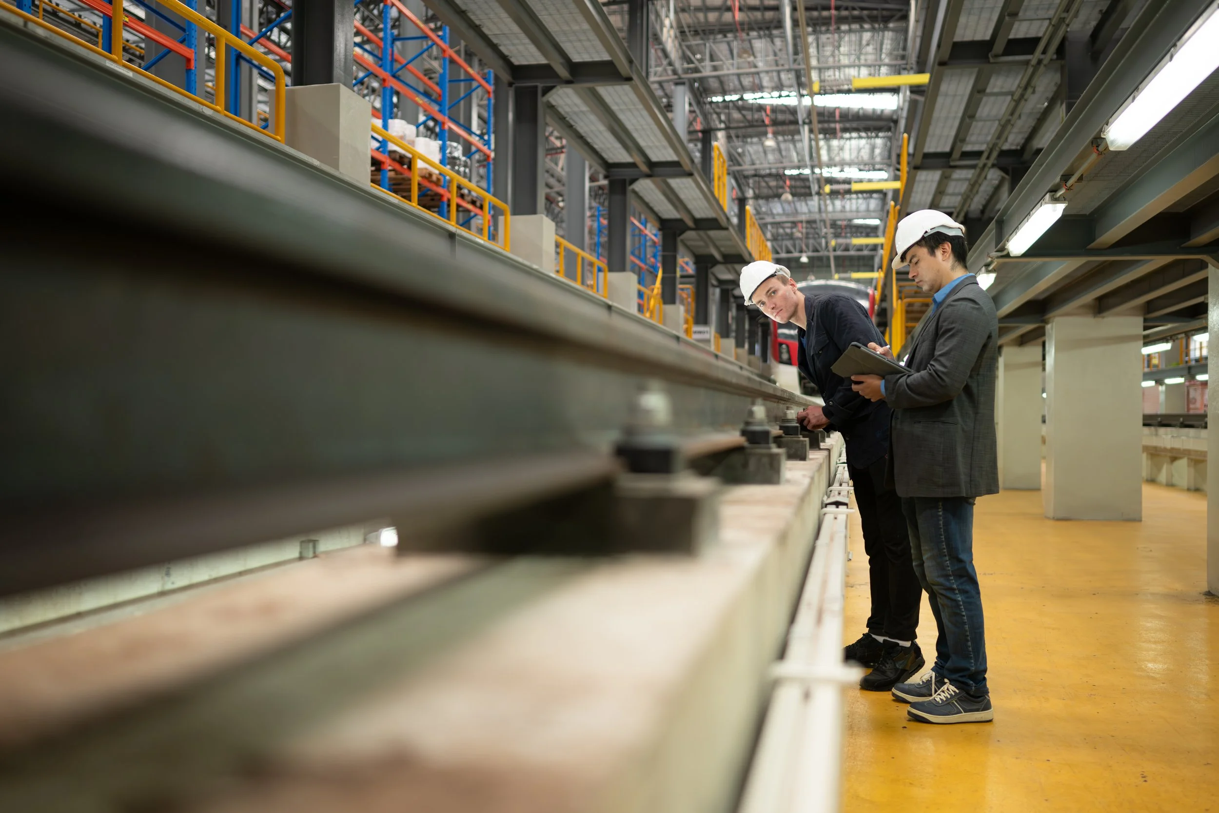 Two men in safety hard hats inspect a rail tie in a factory.