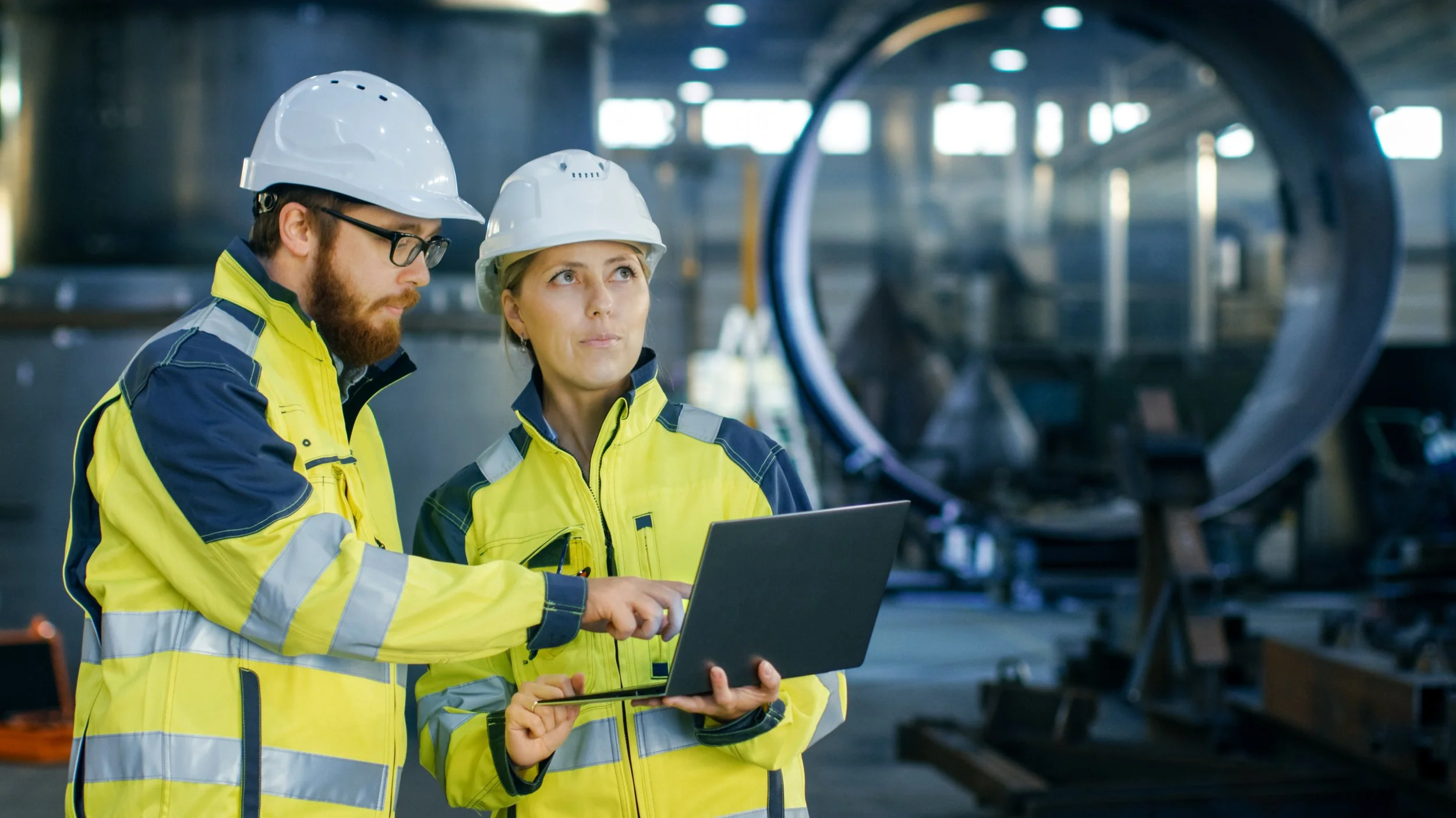 A man and woman in safety gear look at a laptop in an industrial setting.