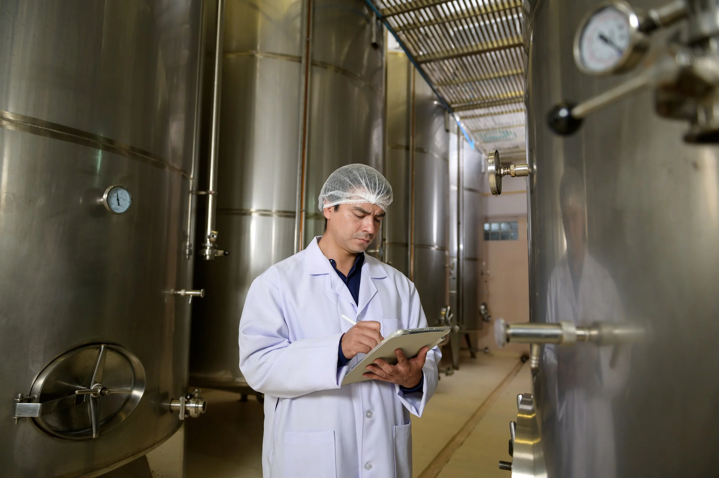 A worker wearing a labcoat and hairnet reviews a tablet while inspecting a wine production plant.