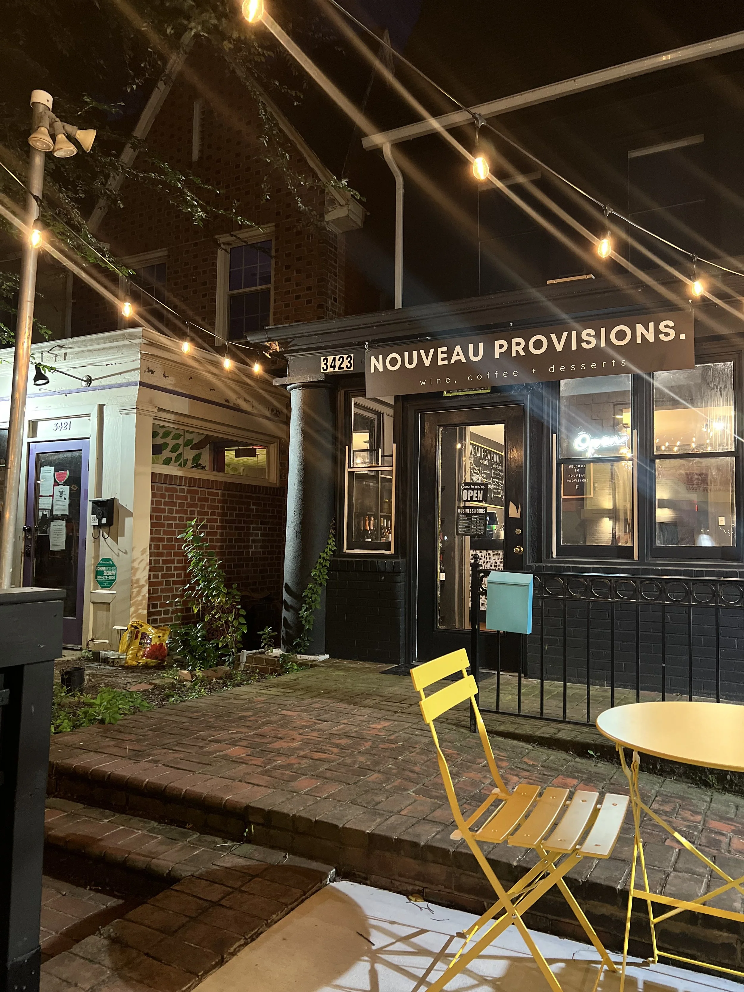 NOUVEAU PROVISIONS in Carytown, Richmond VA, outdoor patio facing Cary St. with string lighting and music. There are yellow metal chairs and a small round table outside, brick sidewalk, and a brick building with windows in the background.