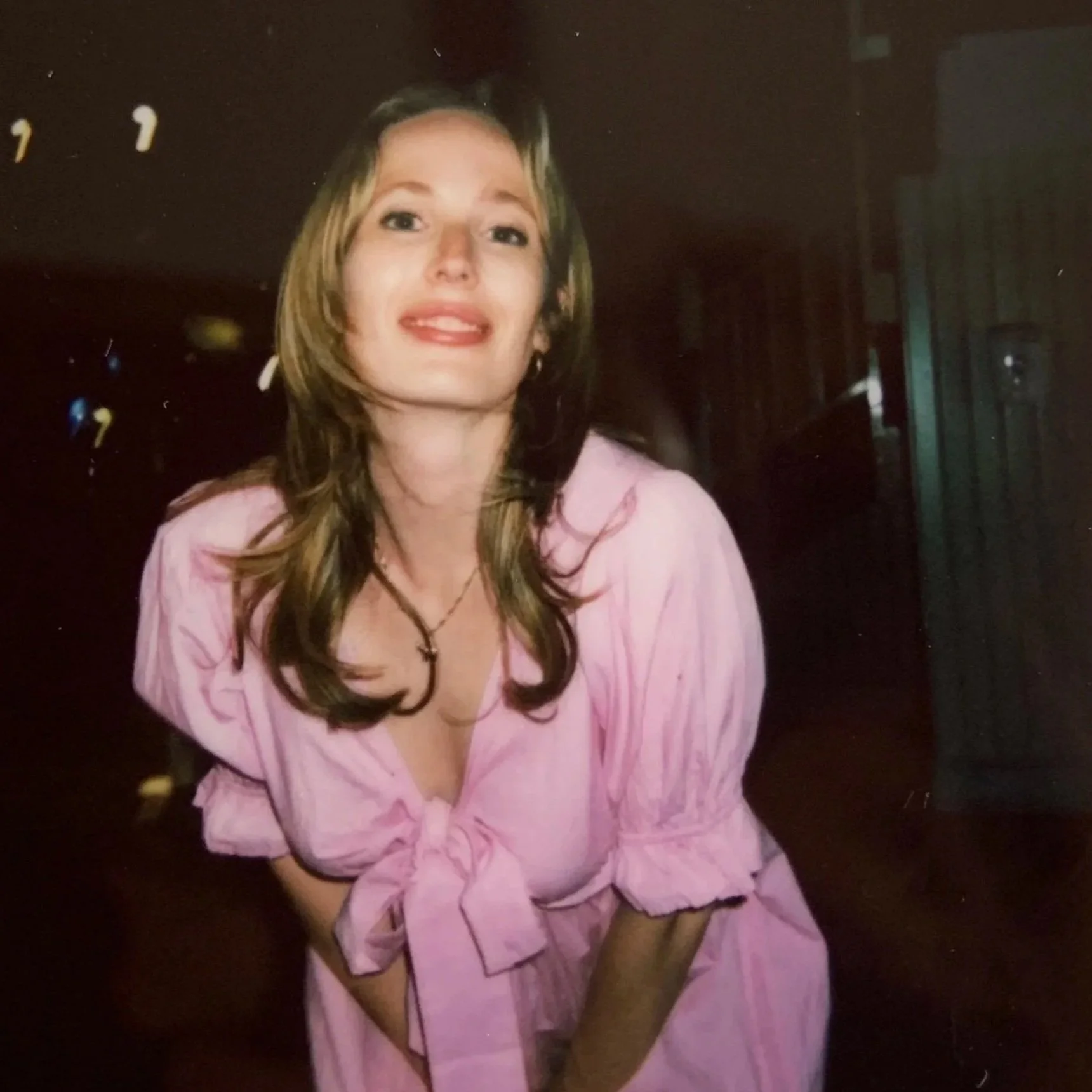 A woman with long brown hair wearing a pink dress with a bow at the waist, smiling and leaning forward.