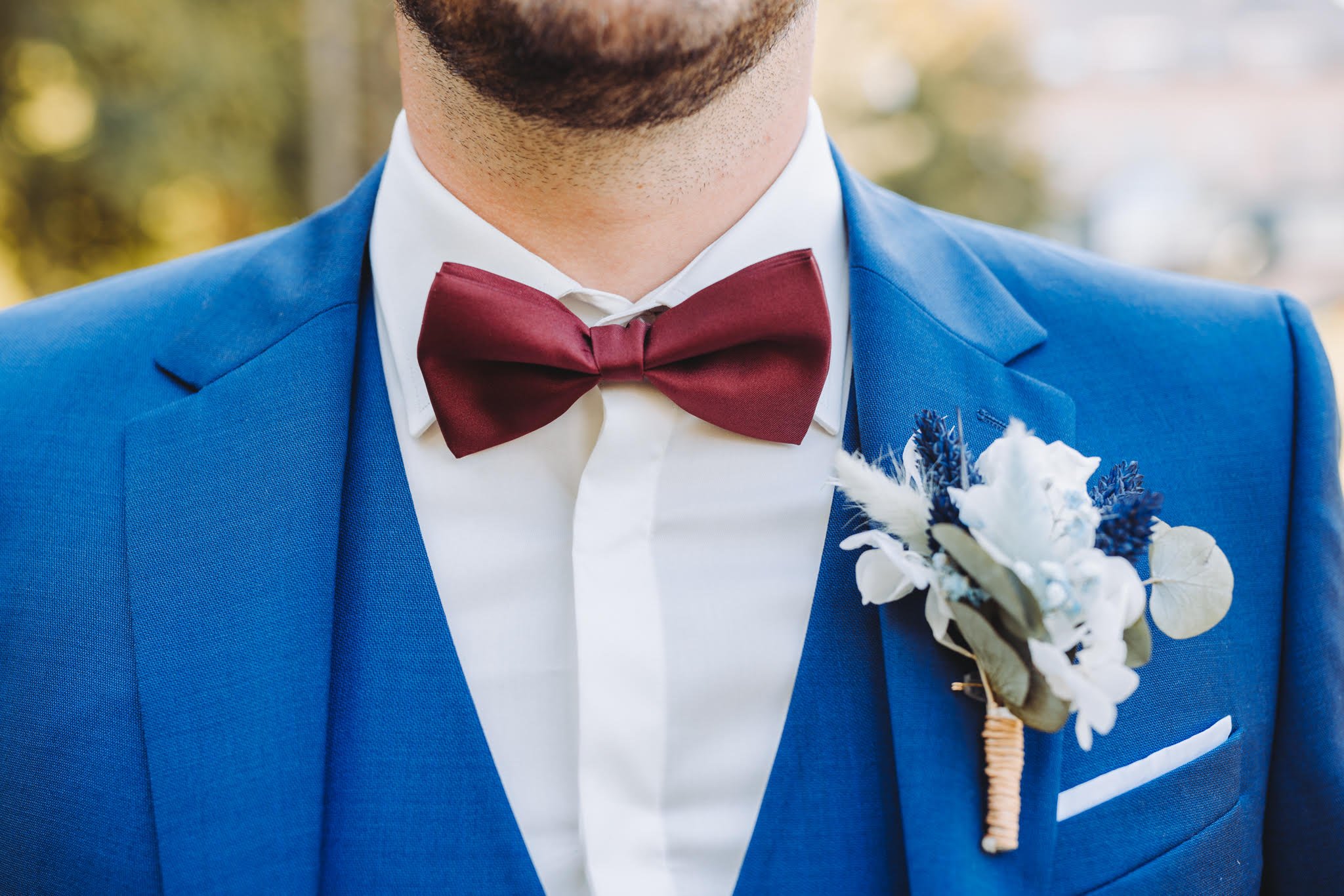 Homme portant un costume bleu avec une chemise blanche, un nœud papillon rouge et une boutonnière avec des fleurs blanches et violettes.