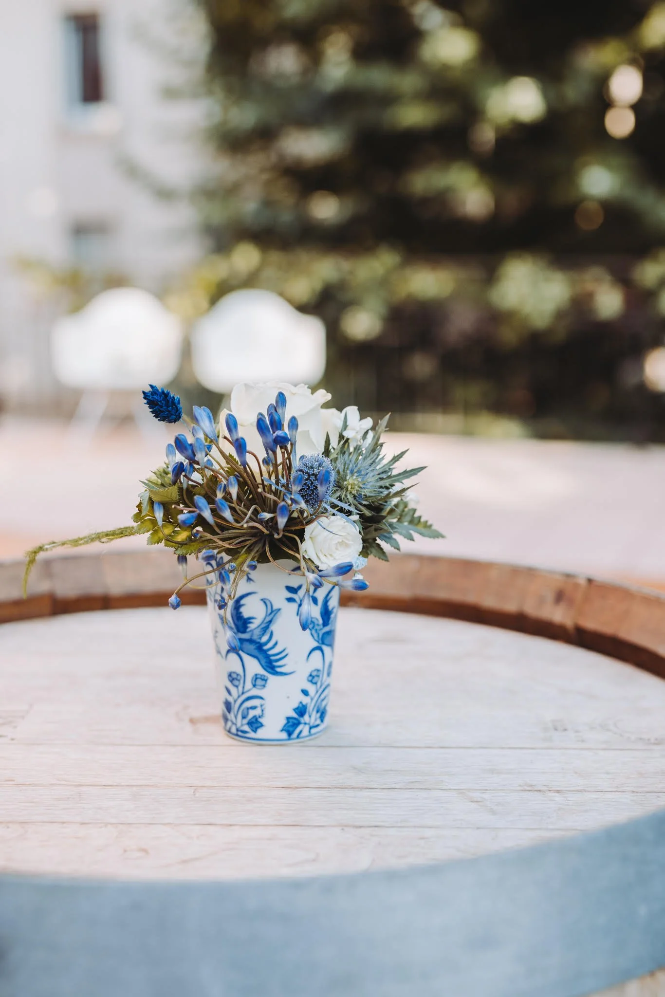 Un arrangement floral dans un vase blanc et bleu avec un motif de fleurs, placé sur une table en bois, avec un fond flou d'arbre et de nature.