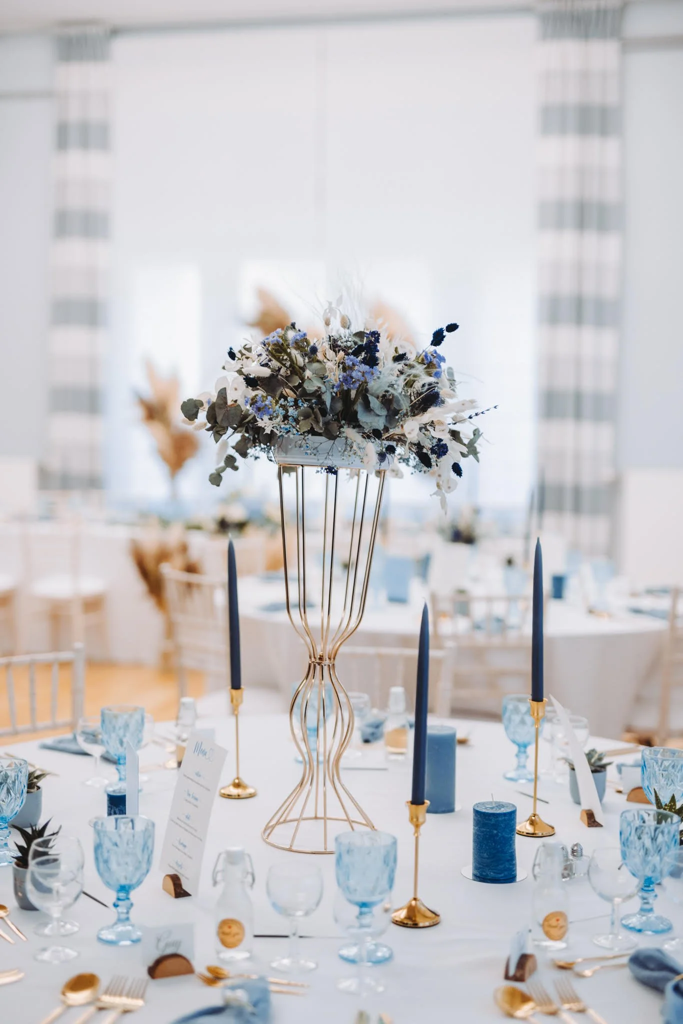 Centre de table avec un vase en forme de cage dorée contenant un bouquet de fleurs blanches et bleues, candelabras avec des bougies bleues et dorées, décoration de table élégante pour un event 