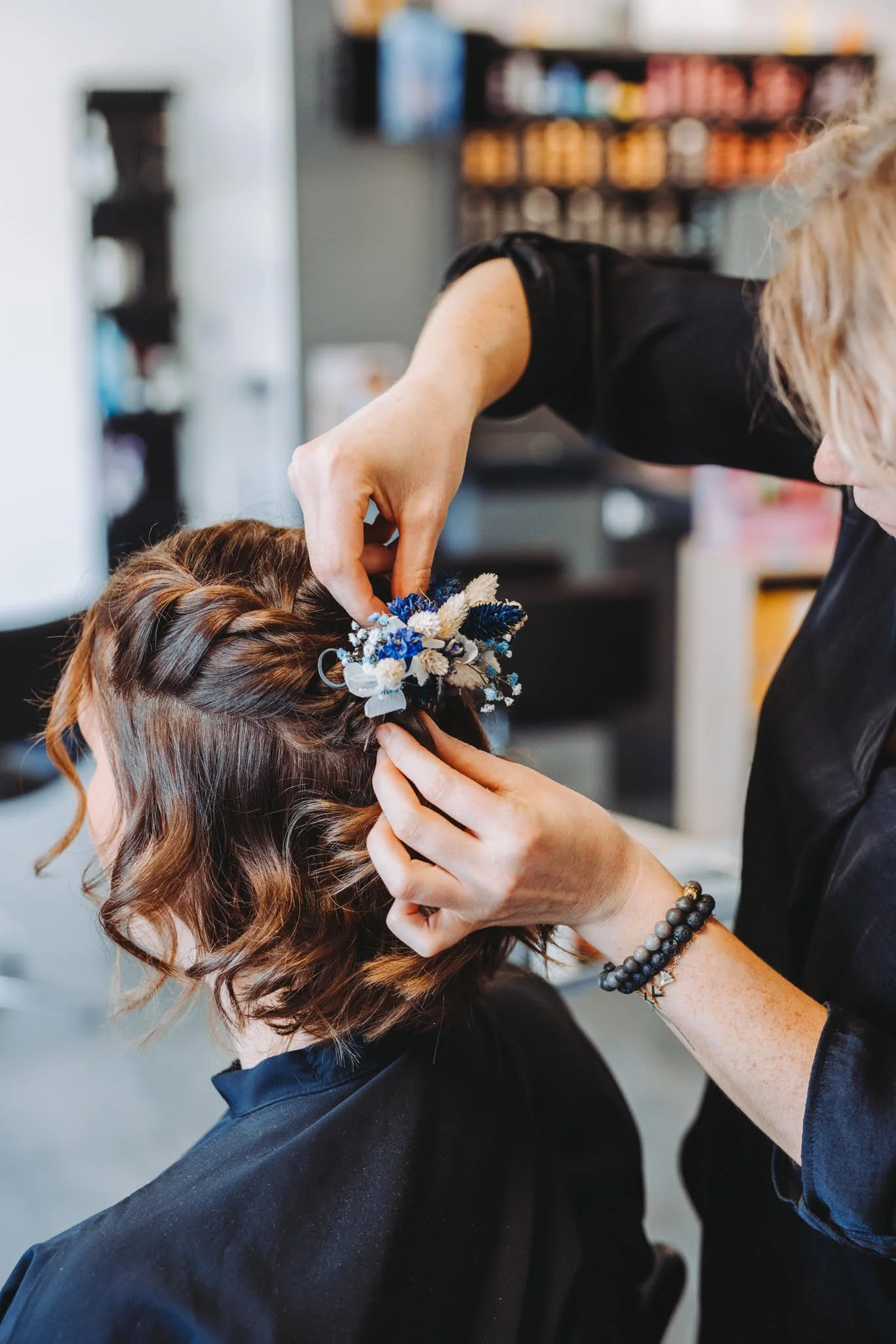 Une femme femme attache une coiffe florale bleue et blanche dans les cheveux d'une autre femme lors d'une séance de coiffure en intérieur.