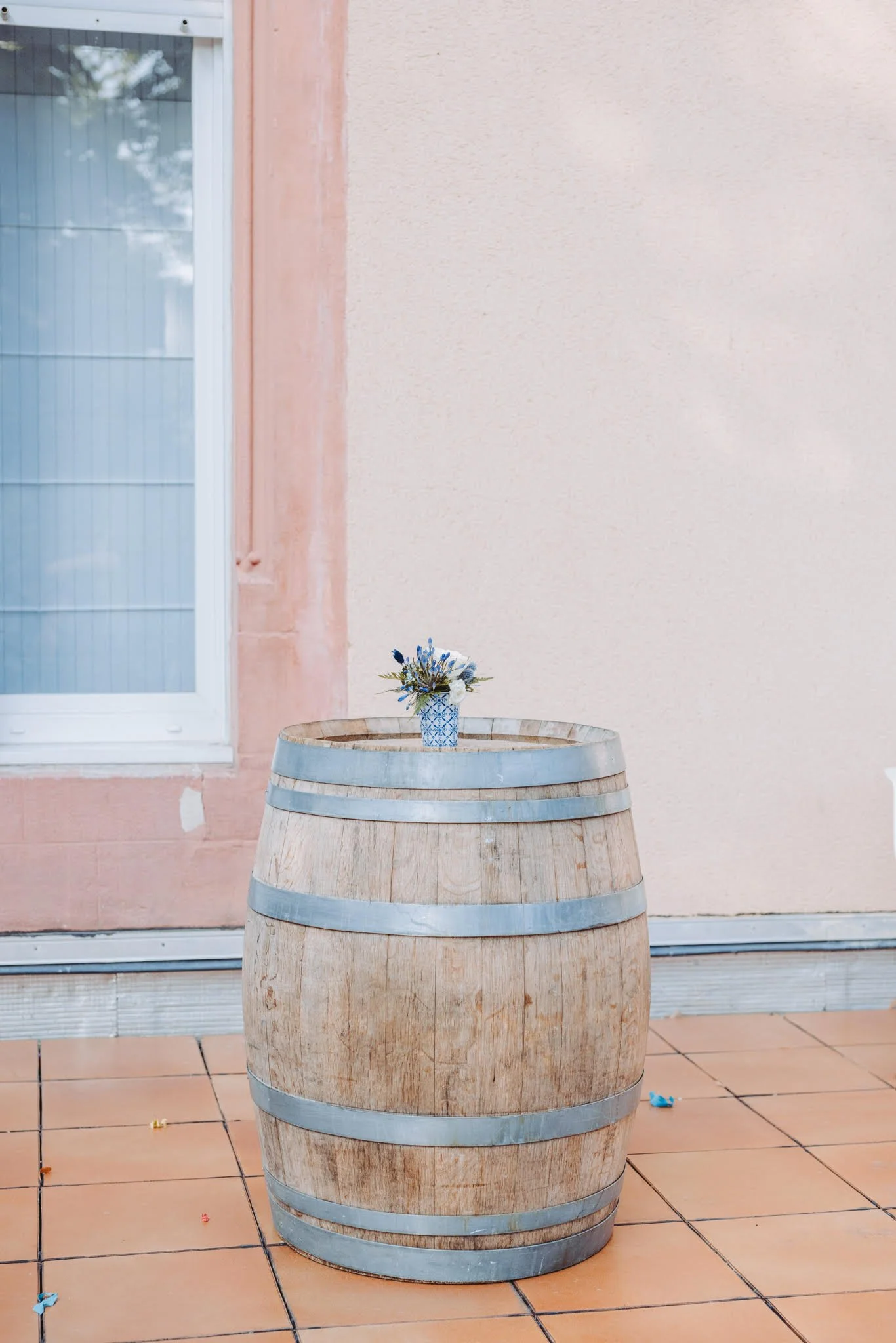 Une table en tonneau en bois posée sur un sol en carreaux, avec un vase de fleurs bleues dessus, à côté d'une fenêtre et d'un mur pastel
