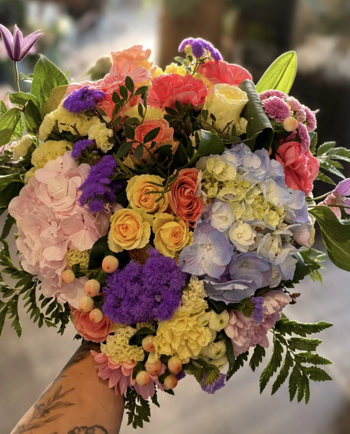 Bouquet de fleurs variées, comprenant des roses jaunes, roses roses, hortensias bleus, fleurs violettes, et feuillage vert, tenu par une personne ayant un tatouage au bras à Florange