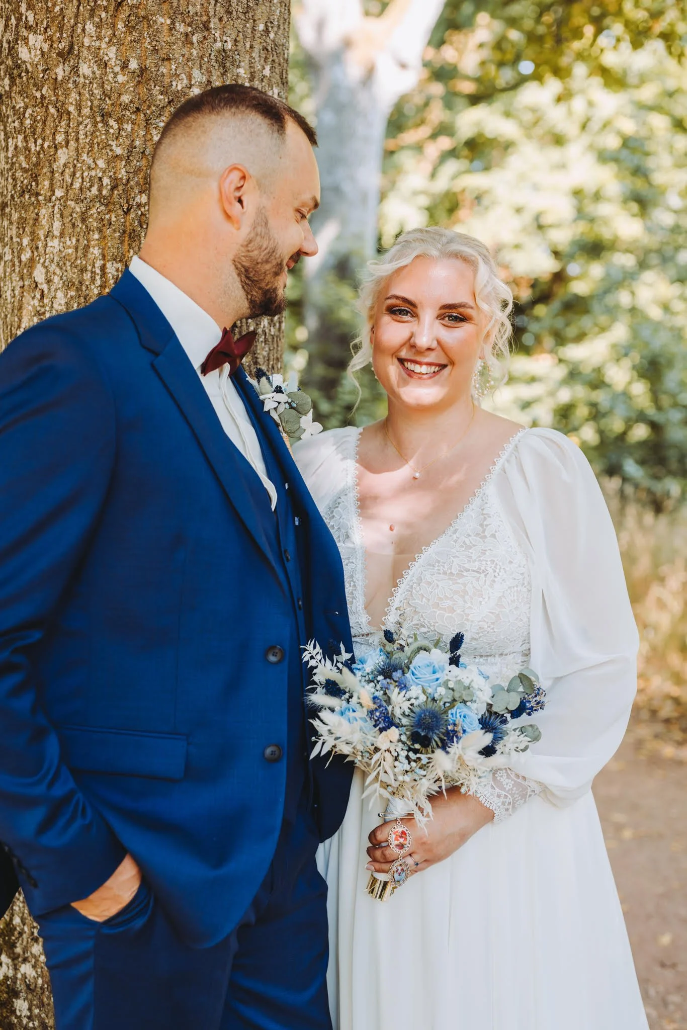 Un couple de mariés souriants dans un parc, la mariée tient un bouquet de fleurs bleues et blanches, l'homme porte un costume bleu foncé, ils se tiennent près d'un arbre, en présence de feuilles vertes et d'une lumière naturelle.