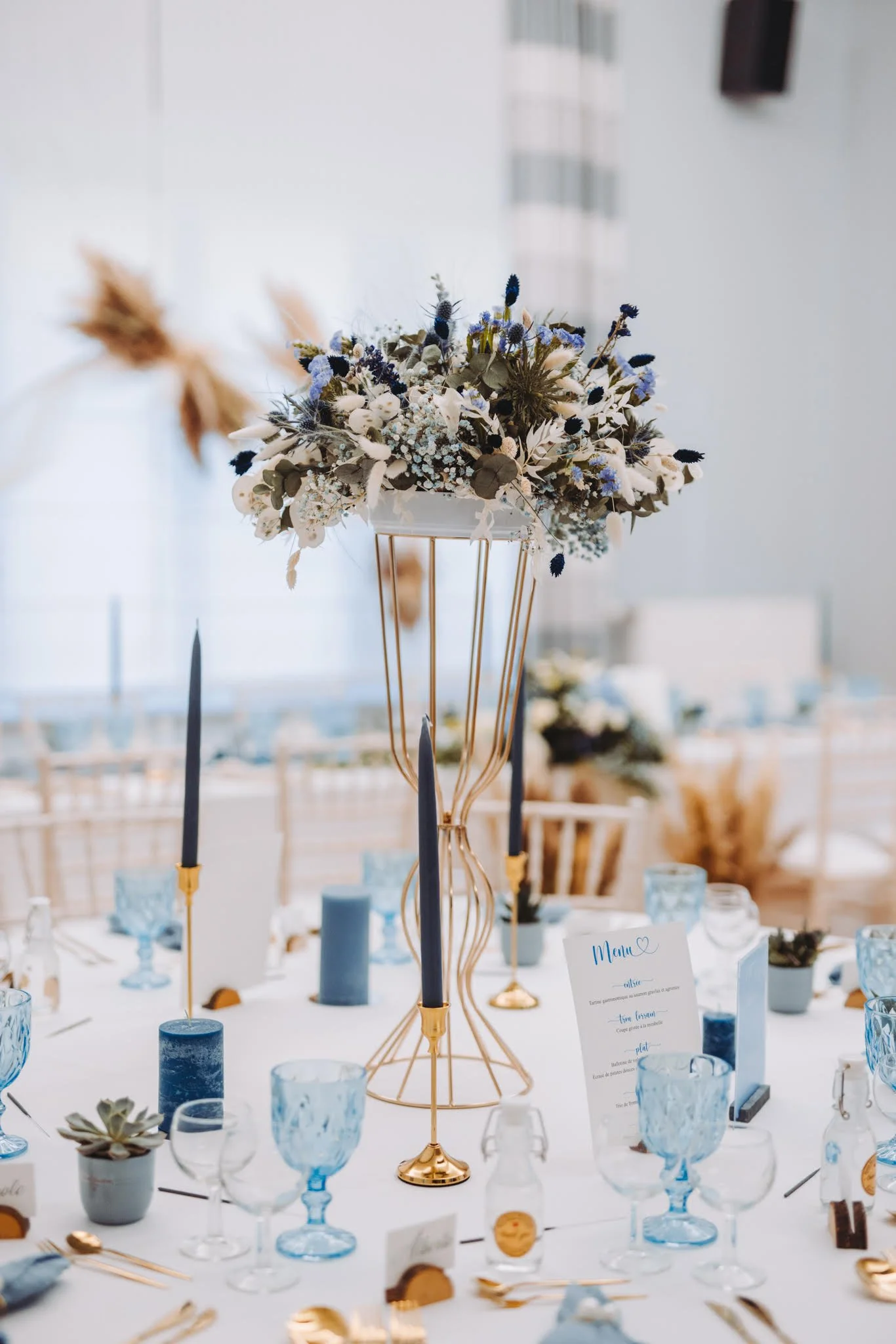 Centre de table avec des fleurs blanches et bleues, flambeaux noirs dans des supports dorés, table de mariage élégante avec des verres bleus et une carte de menu.