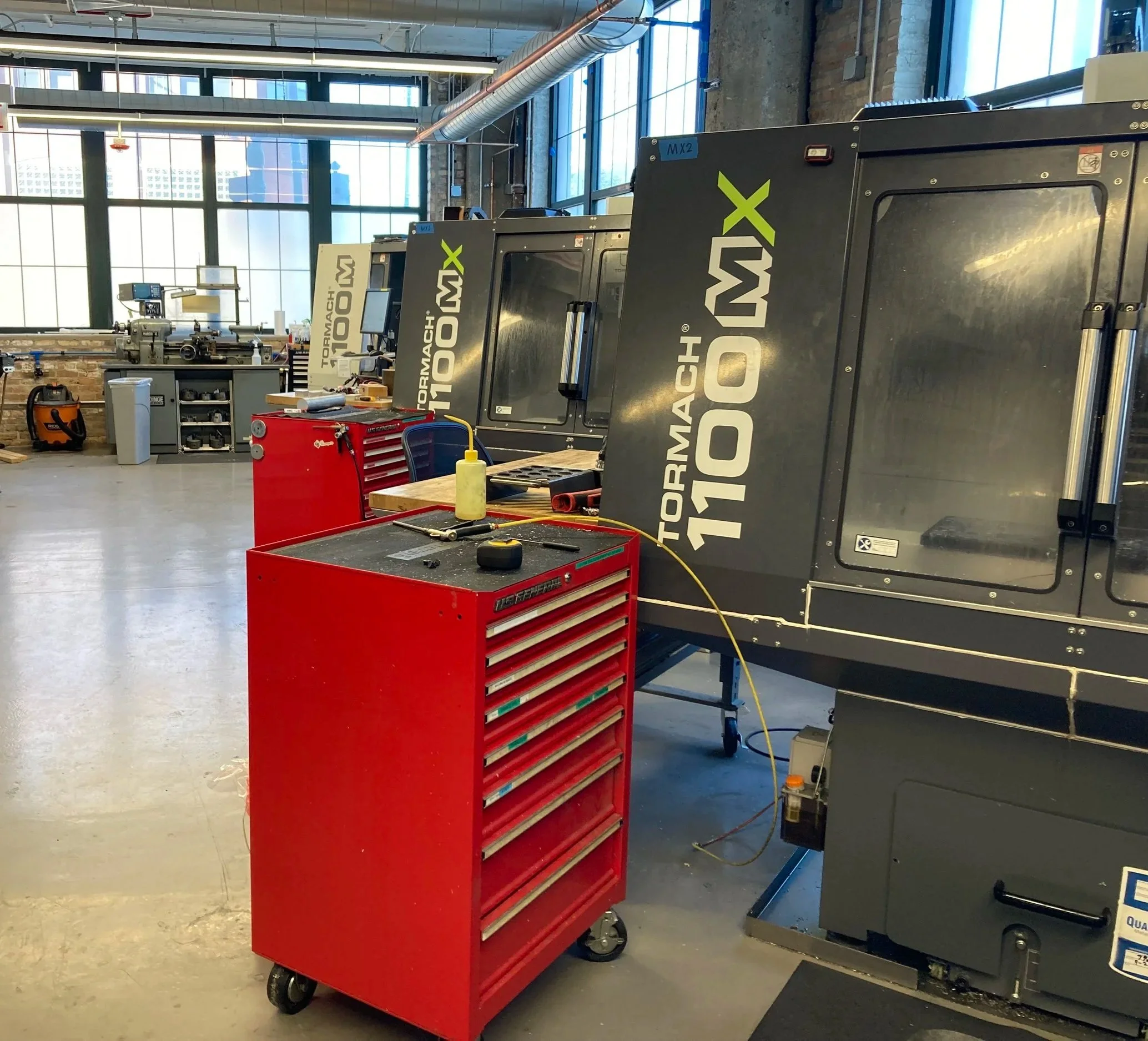 An industrial workshop with large CNC machines labeled Tormach 1100 MX. A red tool chest on wheels with a black top, holding a yellow bottle, a tape measure, and tools, is positioned in front of the machines. The workshop has large windows allowing natural light and visible ductwork and piping on the ceiling.