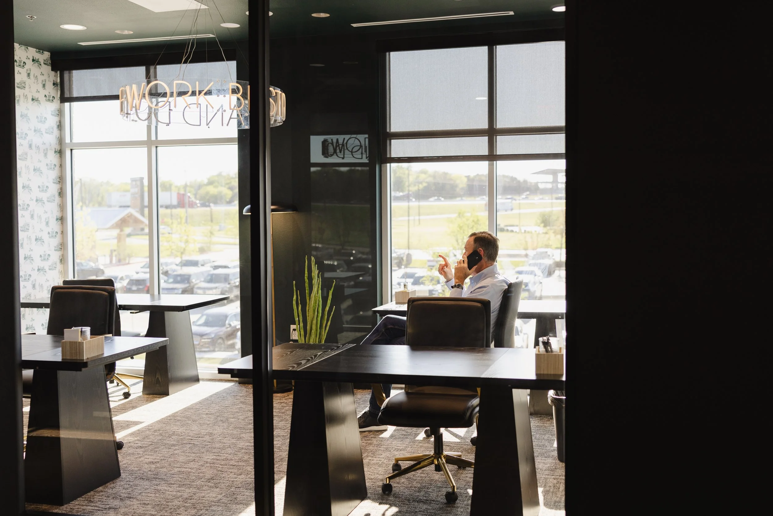 An office space with large windows, some with blinds, showing a parking lot and greenery outside. Inside, a man is seated at a desk, talking on the phone. The decor includes a hanging neon sign that reads 'WORK BLVD' and patterned wallpaper. There are chairs and tables, a cactus plant, and a carpeted floor.