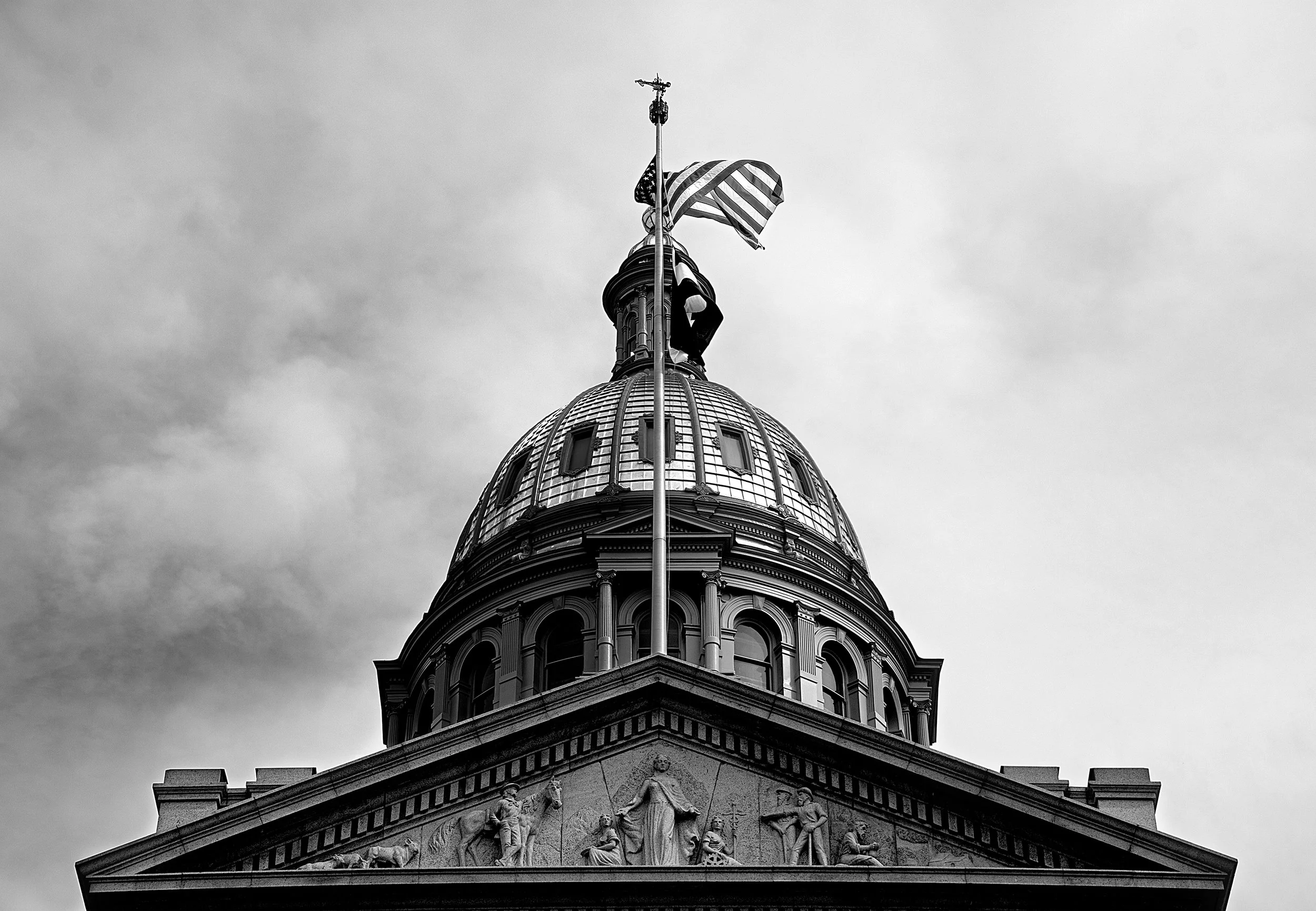 Black and white photo of a domed government building with a flagpole and American flag on top. Architectural details include statues and ornate carvings.