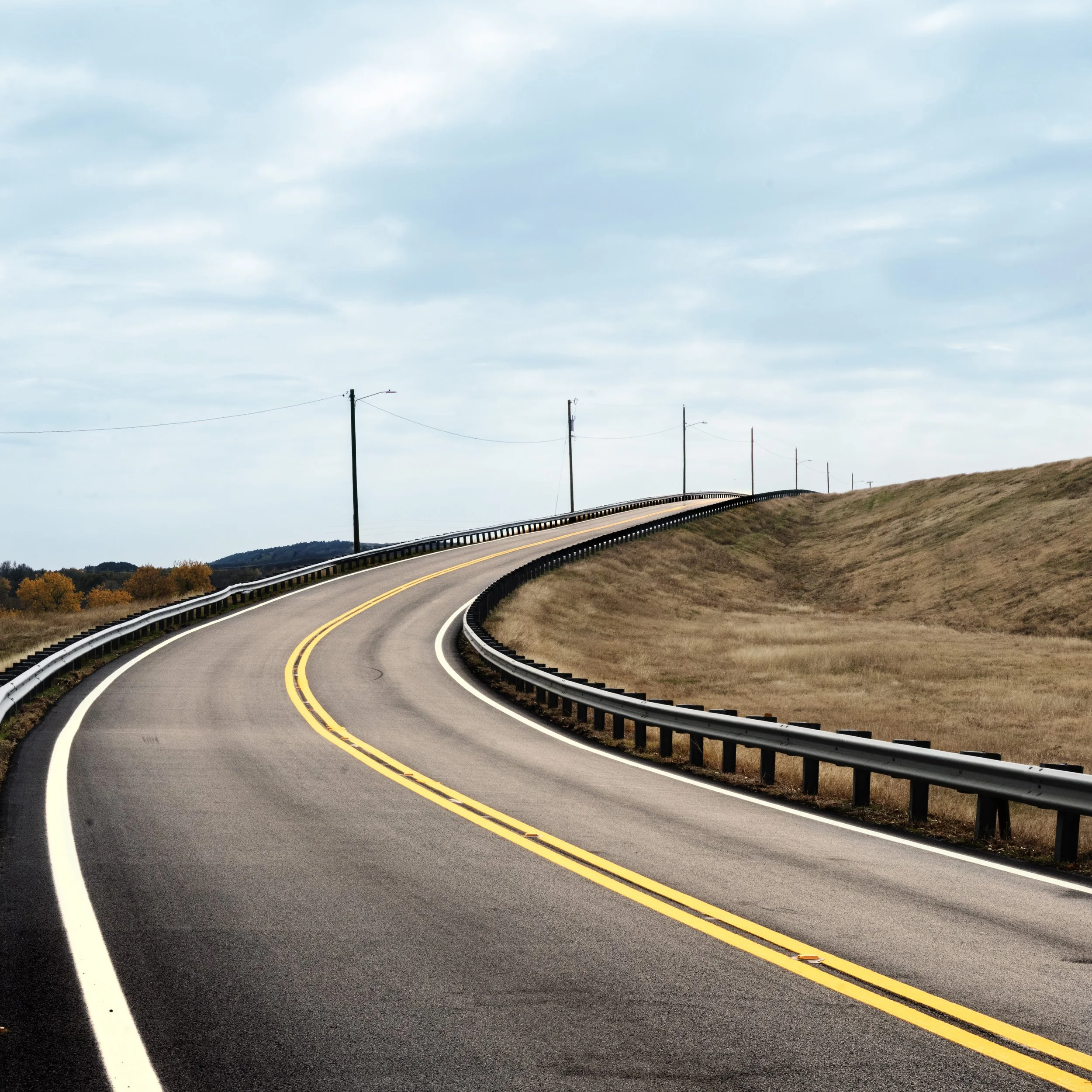 A winding two-lane road with yellow lines, guardrails, and utility poles running alongside, set on a grassy hillside with trees in the distance under a cloudy sky.