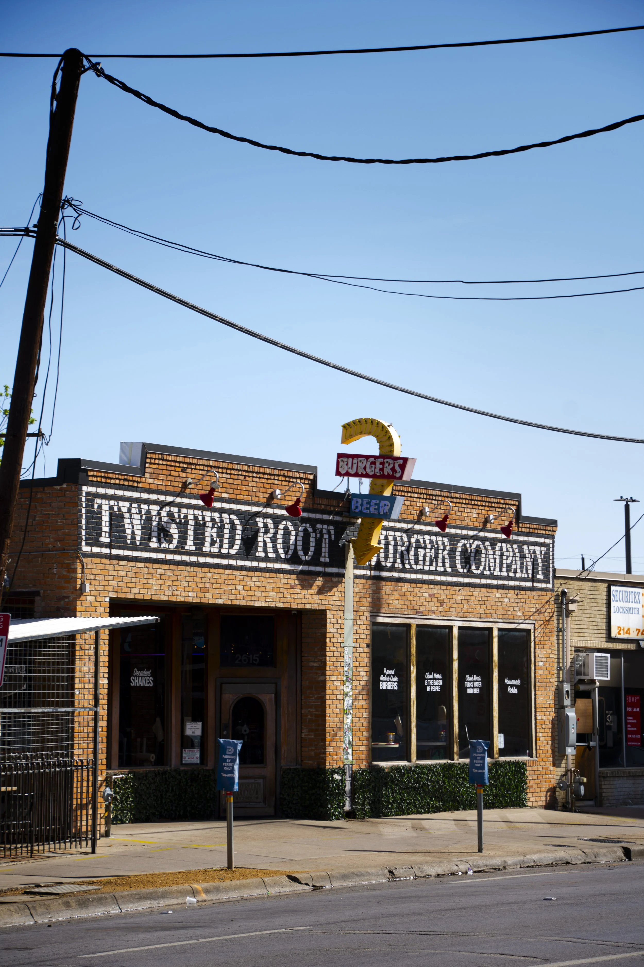 A brick building with a sign promoting TV-tested root beer and burgers. There are large windows, some potted plants outside, and utility poles with wires overhead.